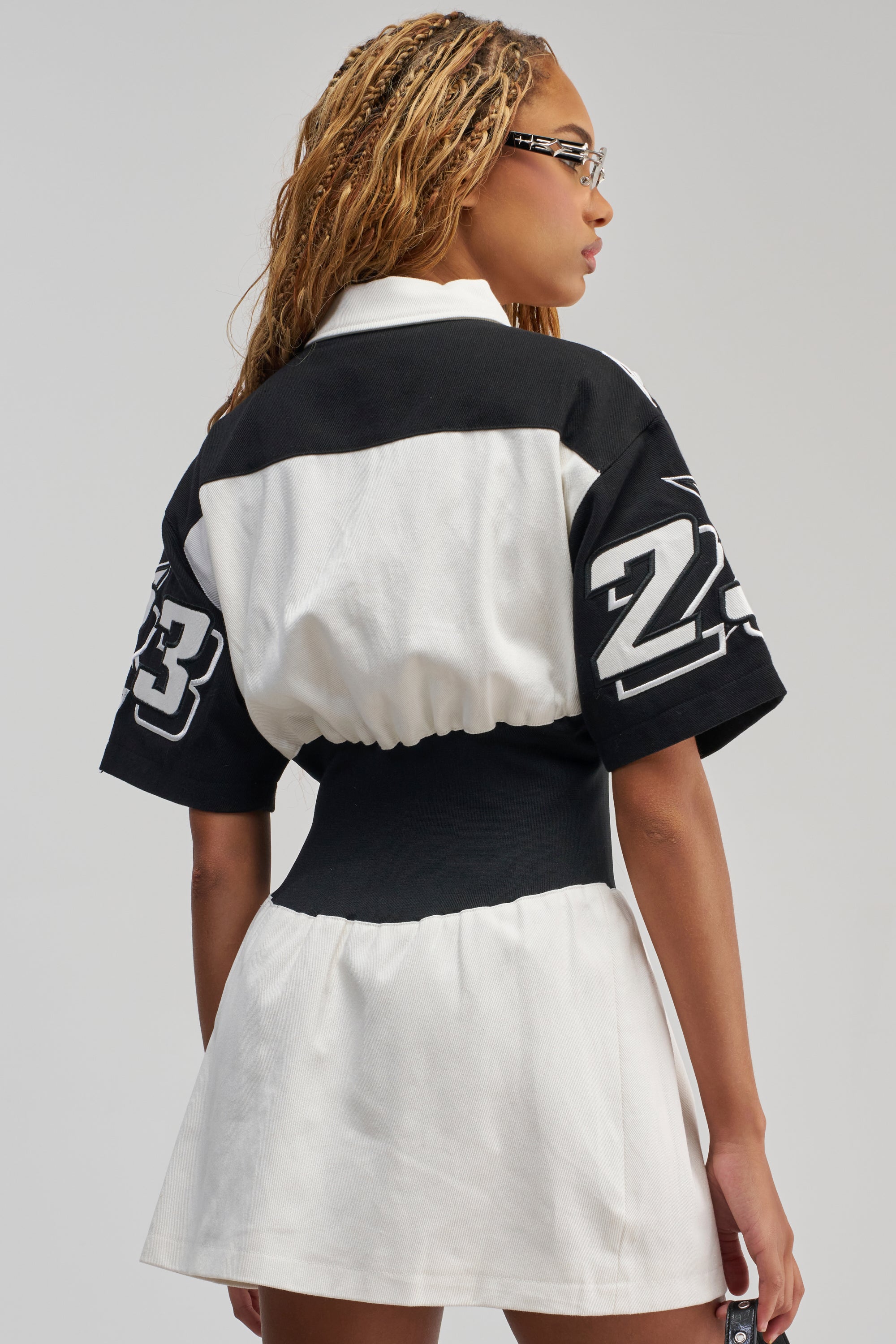 A woman with blonde braids, wearing the OFFICIALLY YOURS COLORBLOCK MINI DRESS in a sporty black and white design with number 23 detailing on the sleeves, stands facing away while turning her head to the side. She accessorizes with glasses and holds a small item in her right hand.