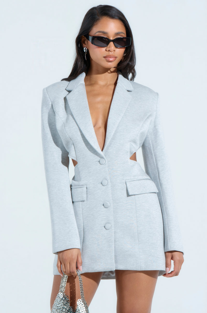 A woman dons the RACHEL SCUBA BLAZER DRESS, a light gray bodycon outfit with cutouts and long sleeves. Featuring a V-neckline, she accessorizes with sunglasses and carries a silver sequin purse against a plain backdrop.