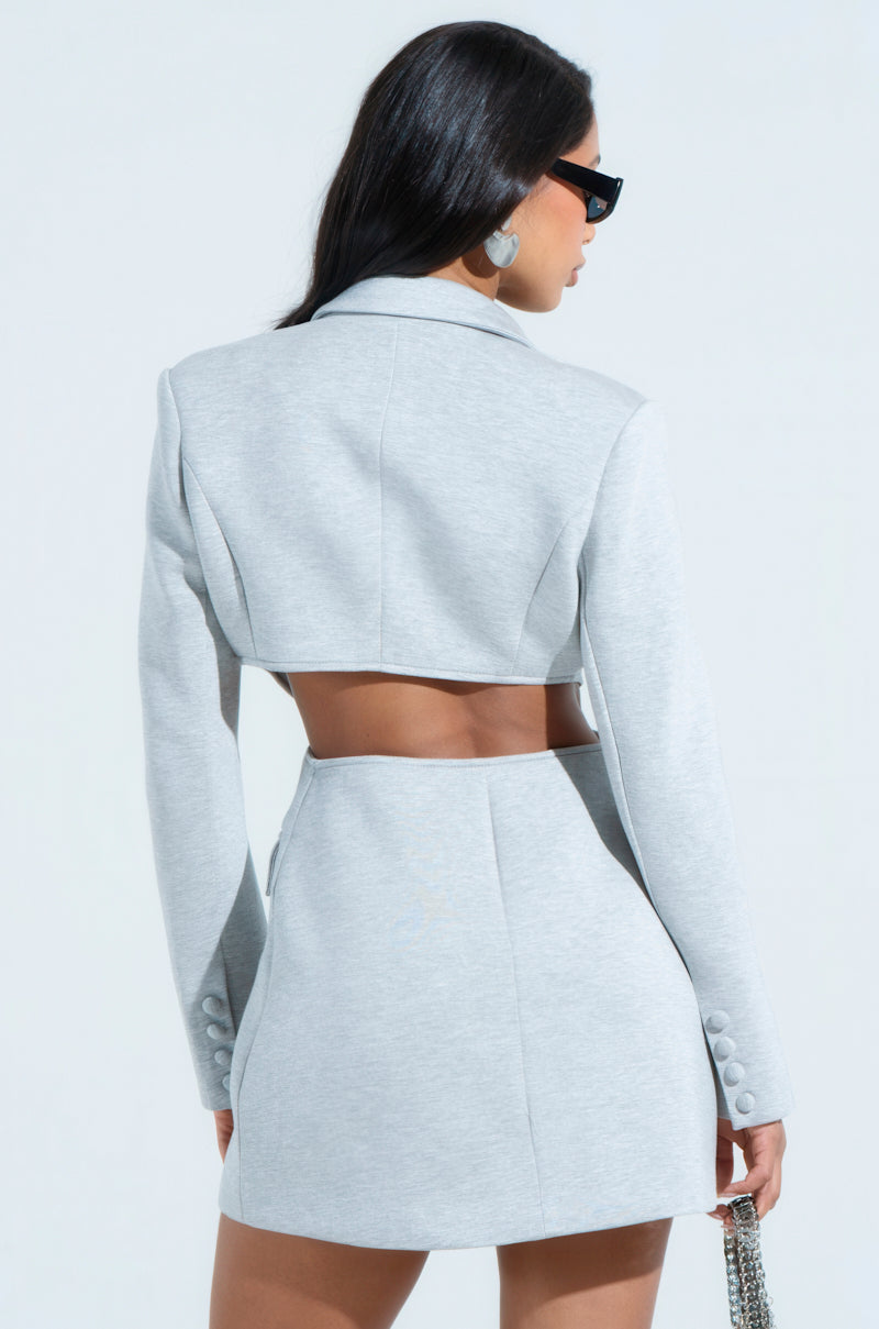 A woman with long dark hair and sunglasses faces away, wearing the RACHEL SCUBA BLAZER DRESS—a stylish, light gray cropped blazer with a V-neckline and high-waisted skirt set. The outfit boasts a cut-out back design and bodycon silhouette while she carries a small handbag.