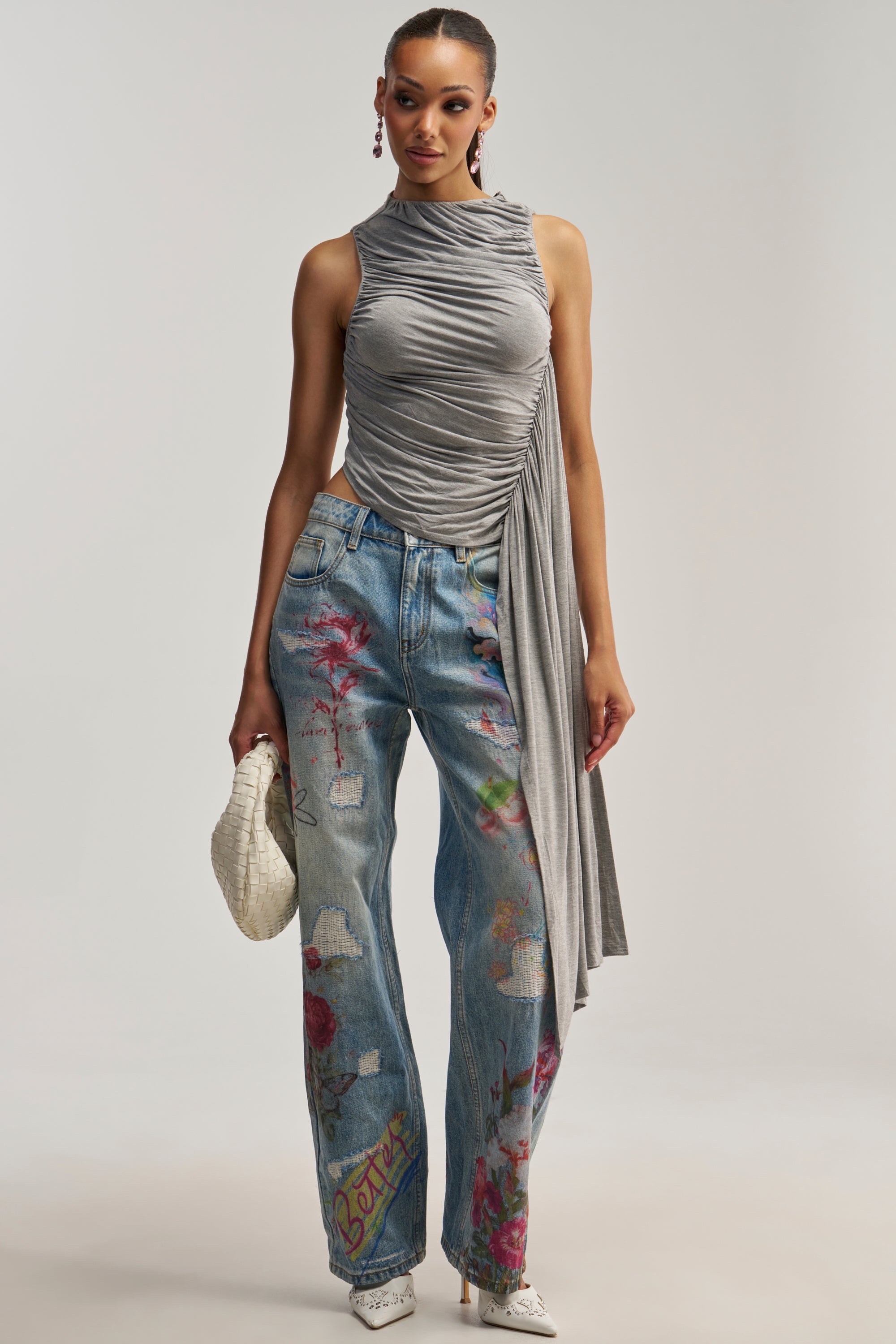 Wearing the KEEPING IT CUTE JERSEY TOP—a sleeveless, ruched jersey piece with an asymmetrical drape—a woman pairs it with painted blue jeans featuring floral designs, white heels, and a white textured clutch. She stands against a plain background, showcasing chic night-out styling with the top’s modern asymmetrical hem elevating her look.
