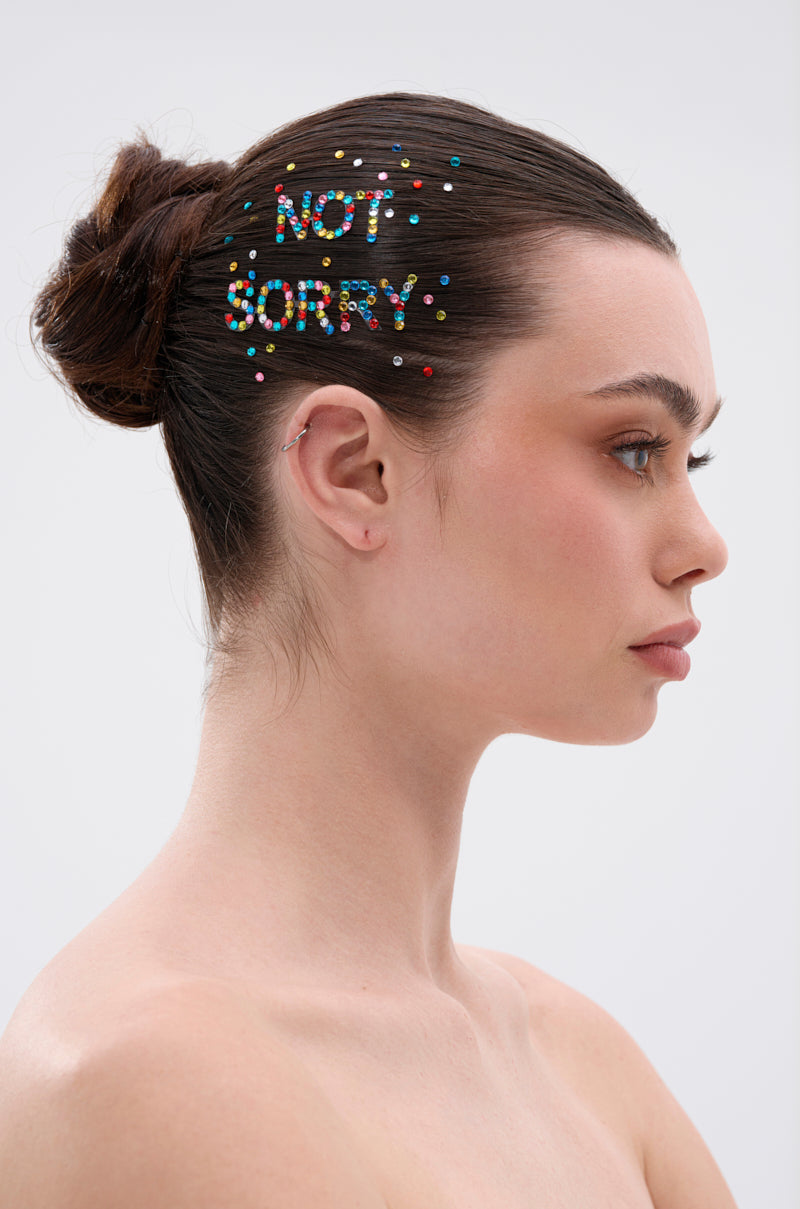 A woman with dark hair in a bun wears NOT SORRY HAIR GEMS that spell “NOT SORRY” on her head. She stands in profile against a plain, light background with a neutral expression. 0