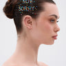 A woman with dark hair in a bun wears NOT SORRY HAIR GEMS that spell “NOT SORRY” on her head. She stands in profile against a plain, light background with a neutral expression.