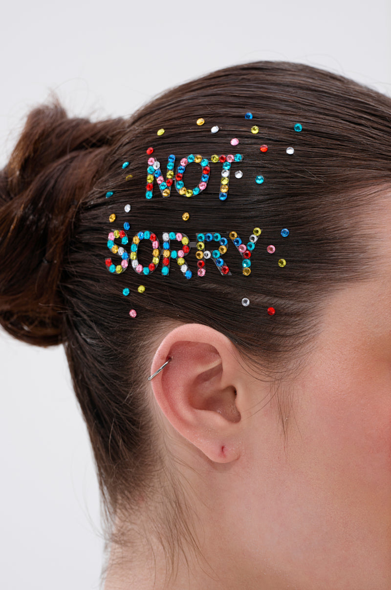 A close-up of brown hair in a bun adorned with NOT SORRY HAIR GEMS—peel-and-stick rhinestones spelling "NOT SORRY" above the ear. The person wears a small hoop earring. 1