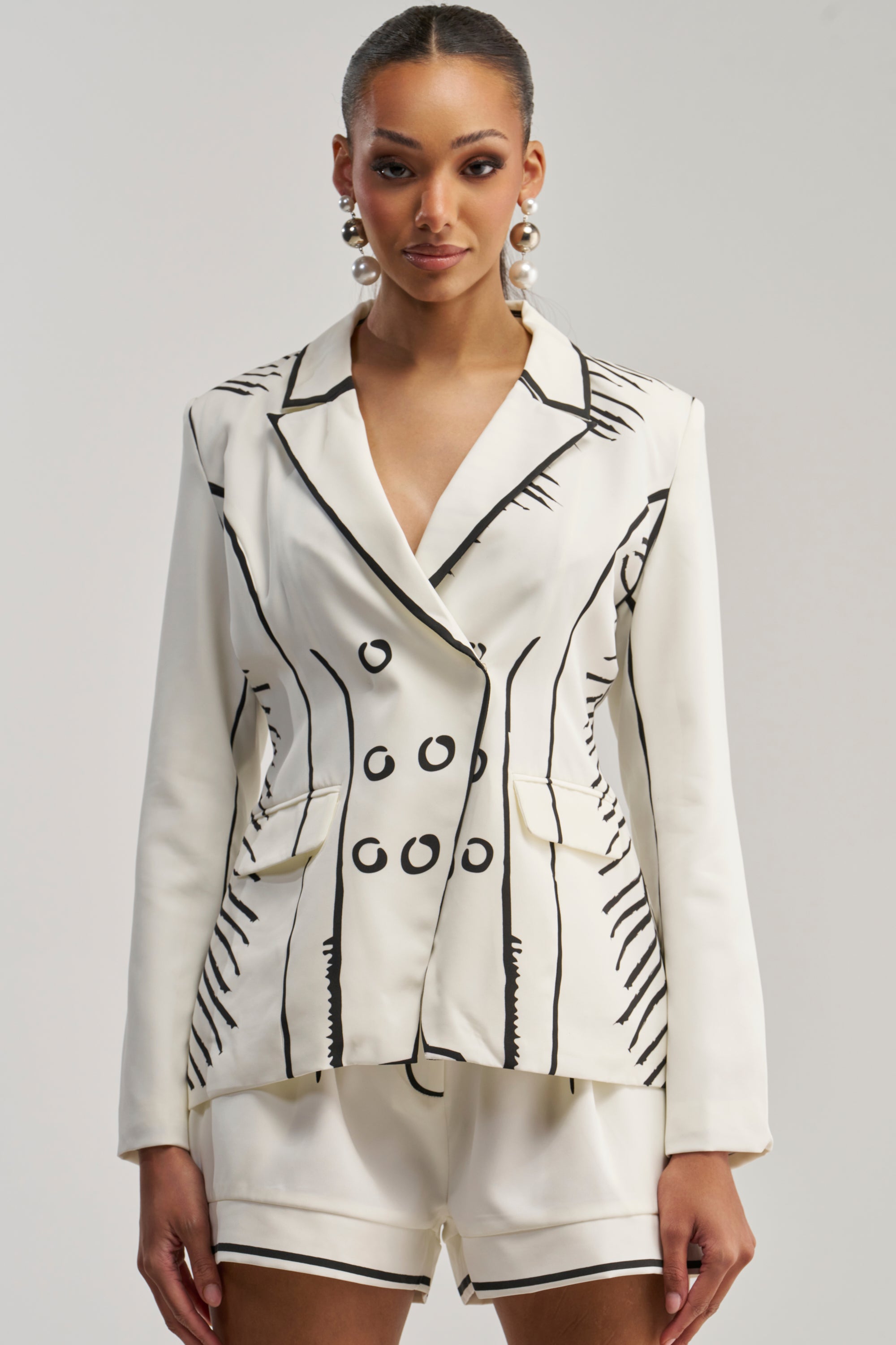 A woman wears the BLANK SPACE BLAZER and matching shorts, each adorned with black sketch-like lines and circles for an illustrated, cartoon-inspired look. She accessorizes with pearl earrings and stands against a plain background, embracing statement styling with her bold outfit.