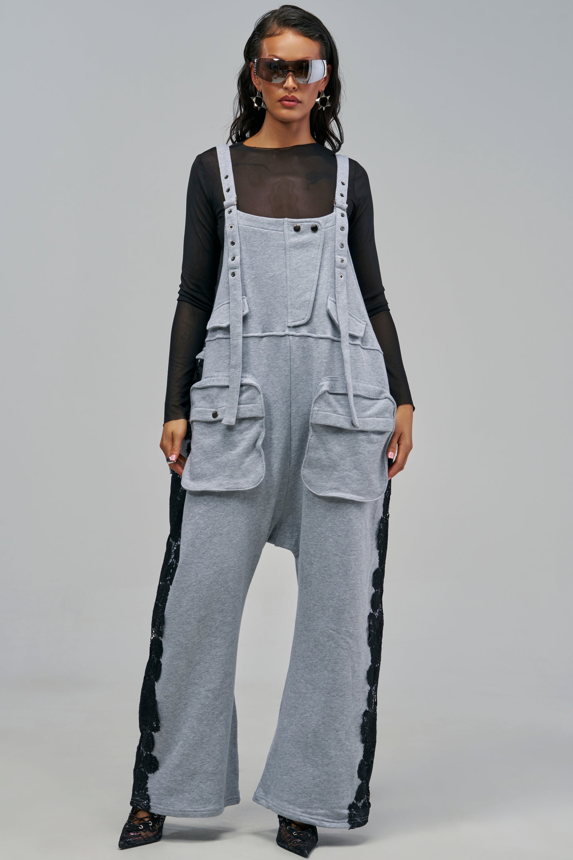 A person wearing the READY IN 5 FRENCH TERRY JUMPSUIT WITH LACE DETAIL in grey, featuring wide-leg utility styling with large cargo pockets and black lace trim, layered over a black long-sleeve mesh top.
