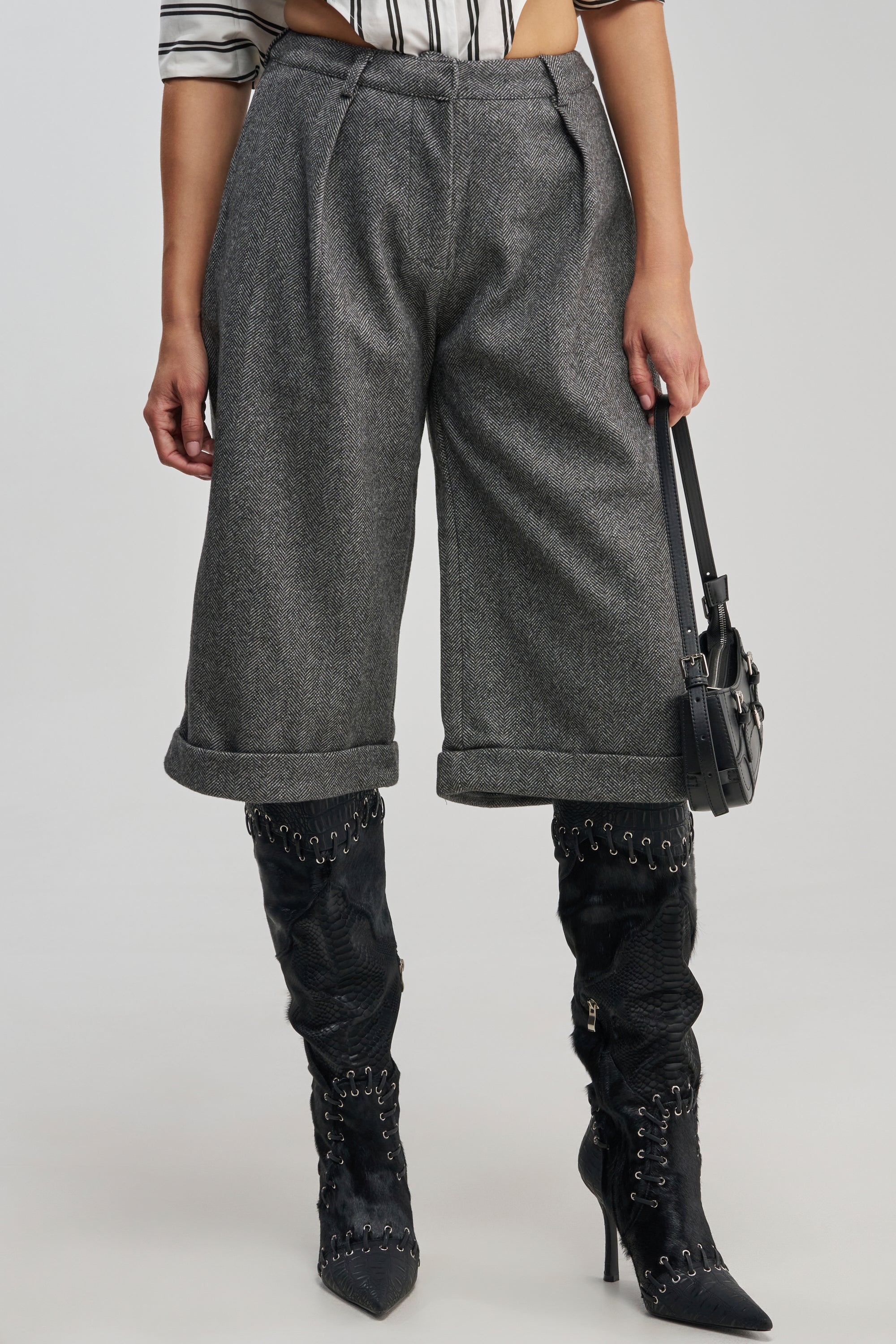 A person wearing the SIREN HIGH RISE TROUSER—gray, wide-legged, cuffed business casual pants—paired with black pointed high-heeled lace-up boots and holding a black handbag. Only the lower torso and legs are visible.