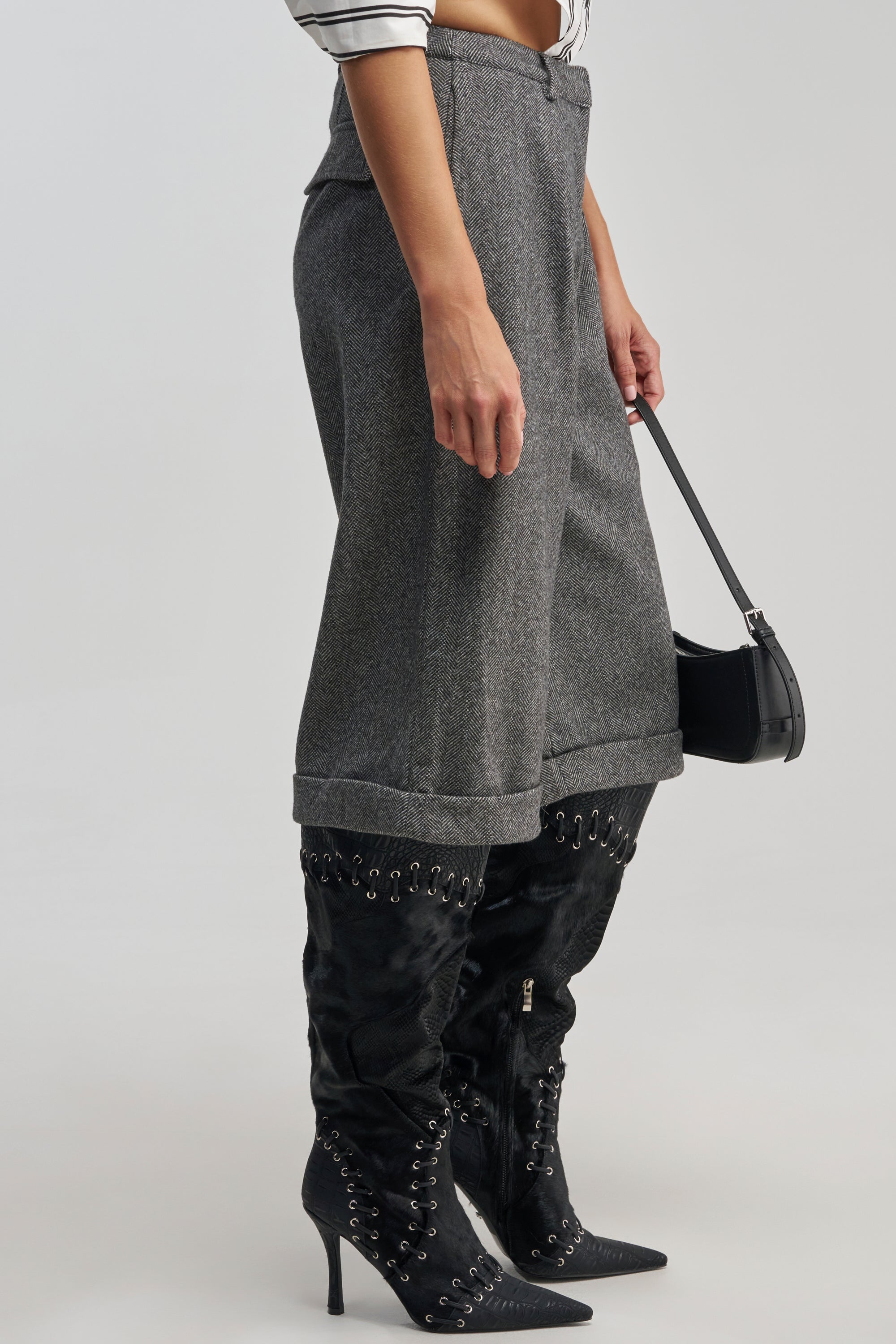 Wearing the SIREN HIGH RISE TROUSER in grey tweed over black lace-up heeled boots and carrying a small black handbag, a person stands against a plain background.