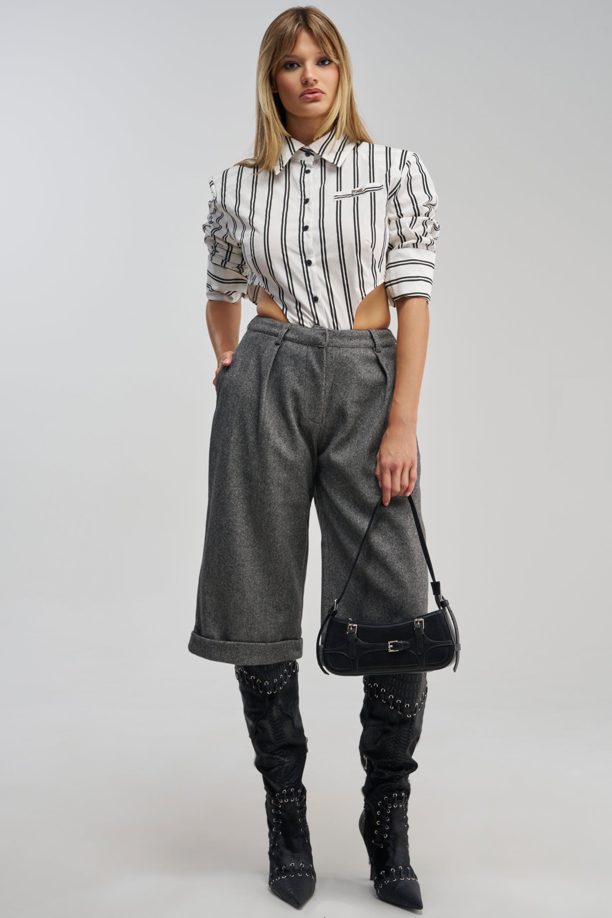 A woman faces forward against a plain background, dressed in the SIREN HIGH RISE TROUSER—a gray cuffed wide-leg pant—paired with a black and white striped button-up shirt featuring side cutouts. She completes the look with studded black knee-high boots and a small black handbag, offering a contemporary take on business casual style through modern lines and bold accents.