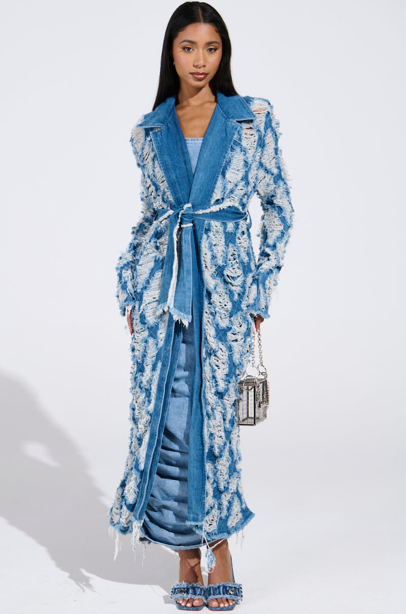 A person poses effortlessly in an ULTRA DISTRESSED DENIM TRENCH over a blue dress with a matching belt against a plain white background, holding a small patterned handbag and wearing denim sandals, completing the stylish look.
