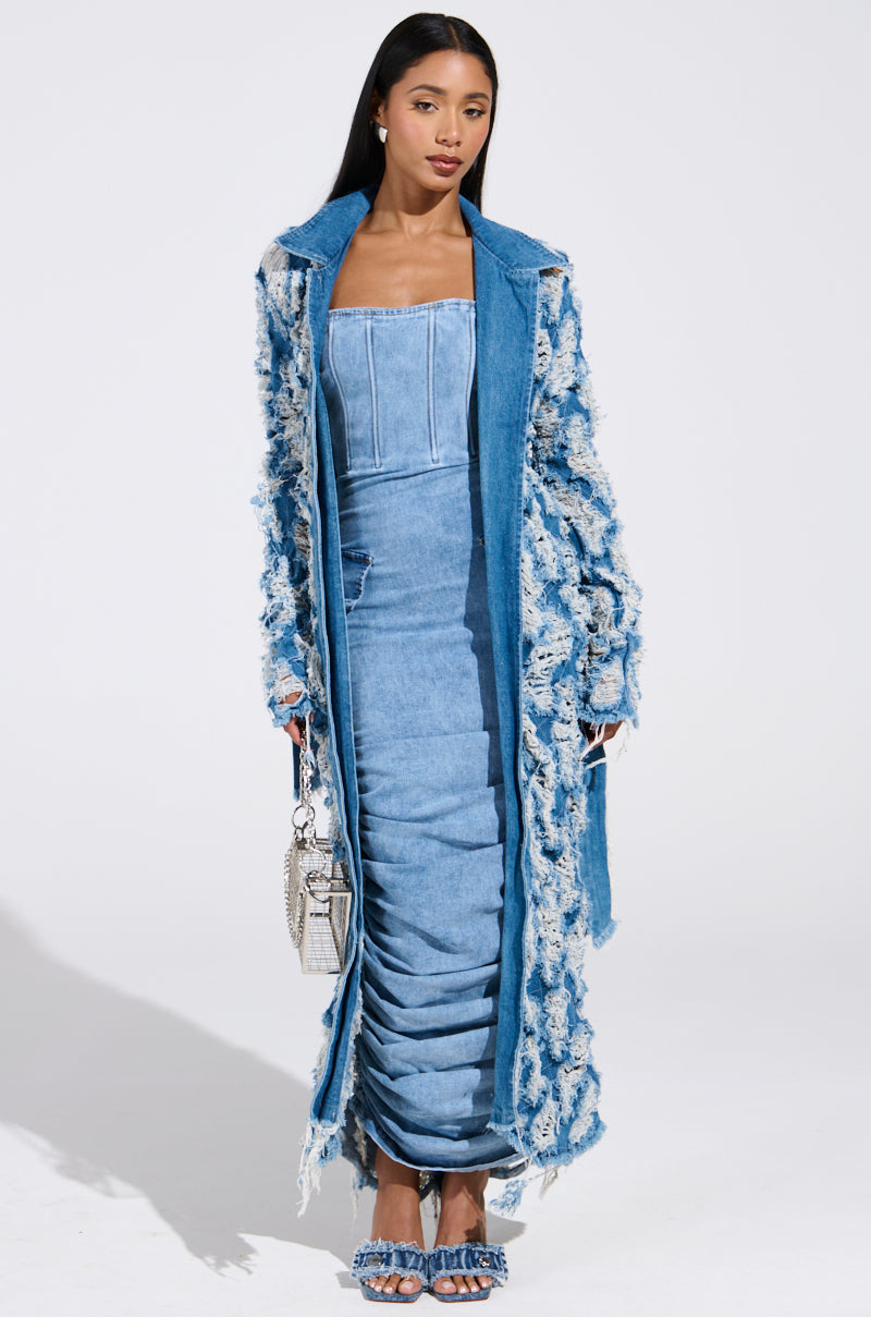 A woman stands against a plain white background wearing the ULTRA DISTRESSED DENIM TRENCH over a strapless, fitted denim dress with ruched detailing. She holds a small silver purse, wears matching stiletto pumps, and her long hair is straight.