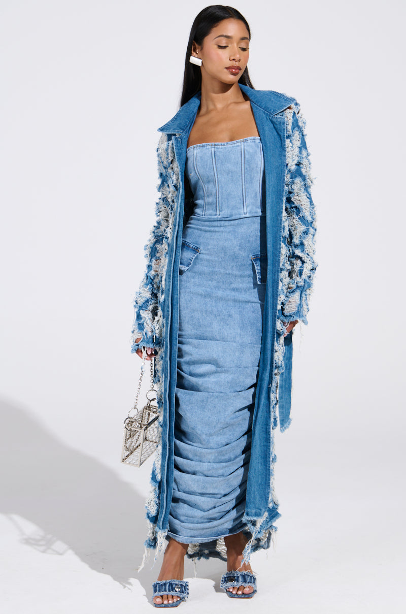 Wearing the ULTRA DISTRESSED DENIM TRENCH over a strapless dress with ruching, they hold a small silver handbag and wear denim stiletto pumps, posed against a plain white backdrop.