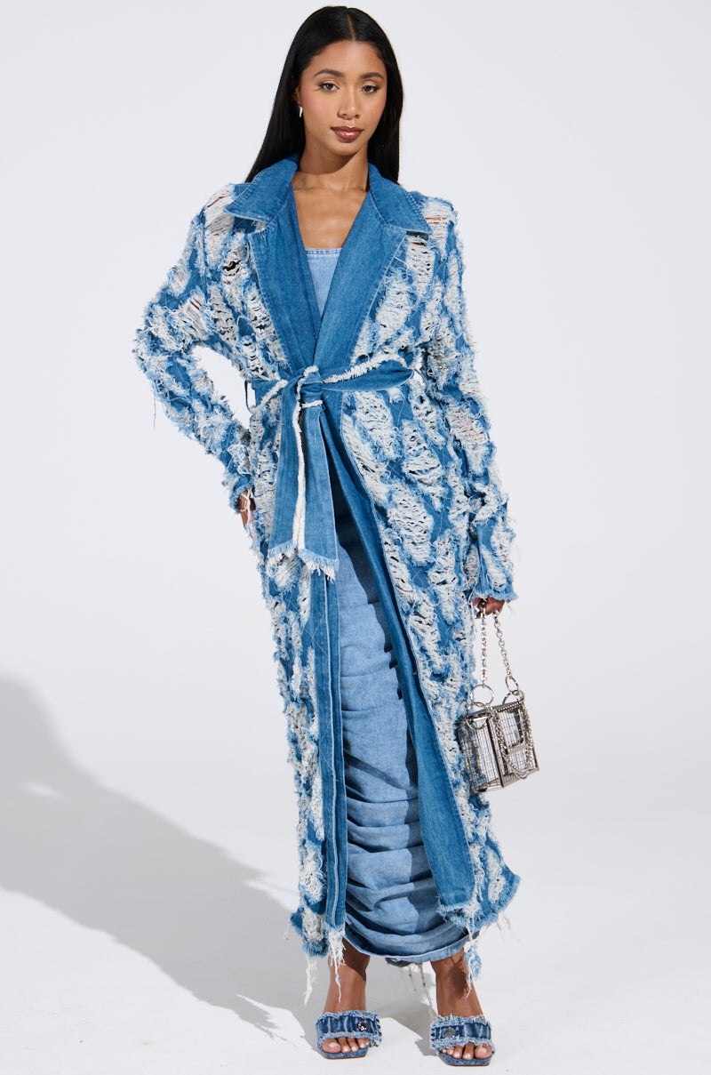 A woman confidently wears the ULTRA DISTRESSED DENIM TRENCH over a light blue midi dress, holding a small silver handbag. Her stiletto pumps and straight, loose hair are highlighted against the plain white background, emphasizing her chic style.