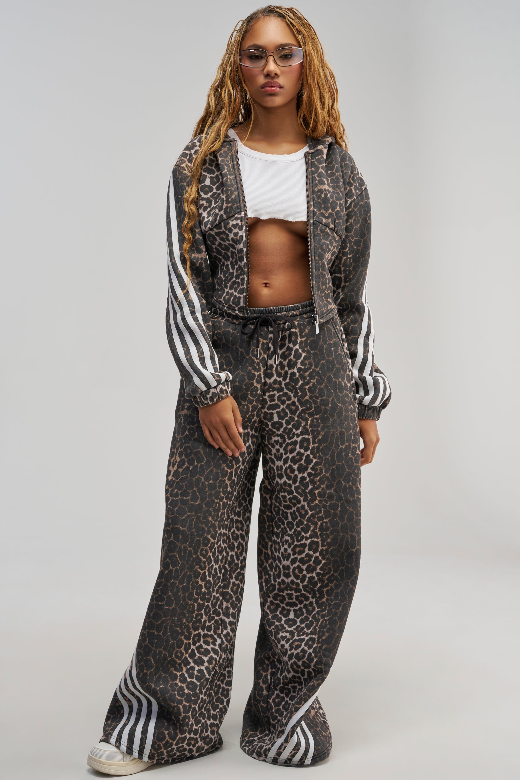 A person with long braids and clear-framed glasses stands confidently against a plain background, wearing the WILD SIDE RELAXED SWEATPANT—a leopard print tracksuit with white stripes—paired with a white crop top. The stylish loungewear set features wide-leg sweatpants for a relaxed yet trendy vibe.