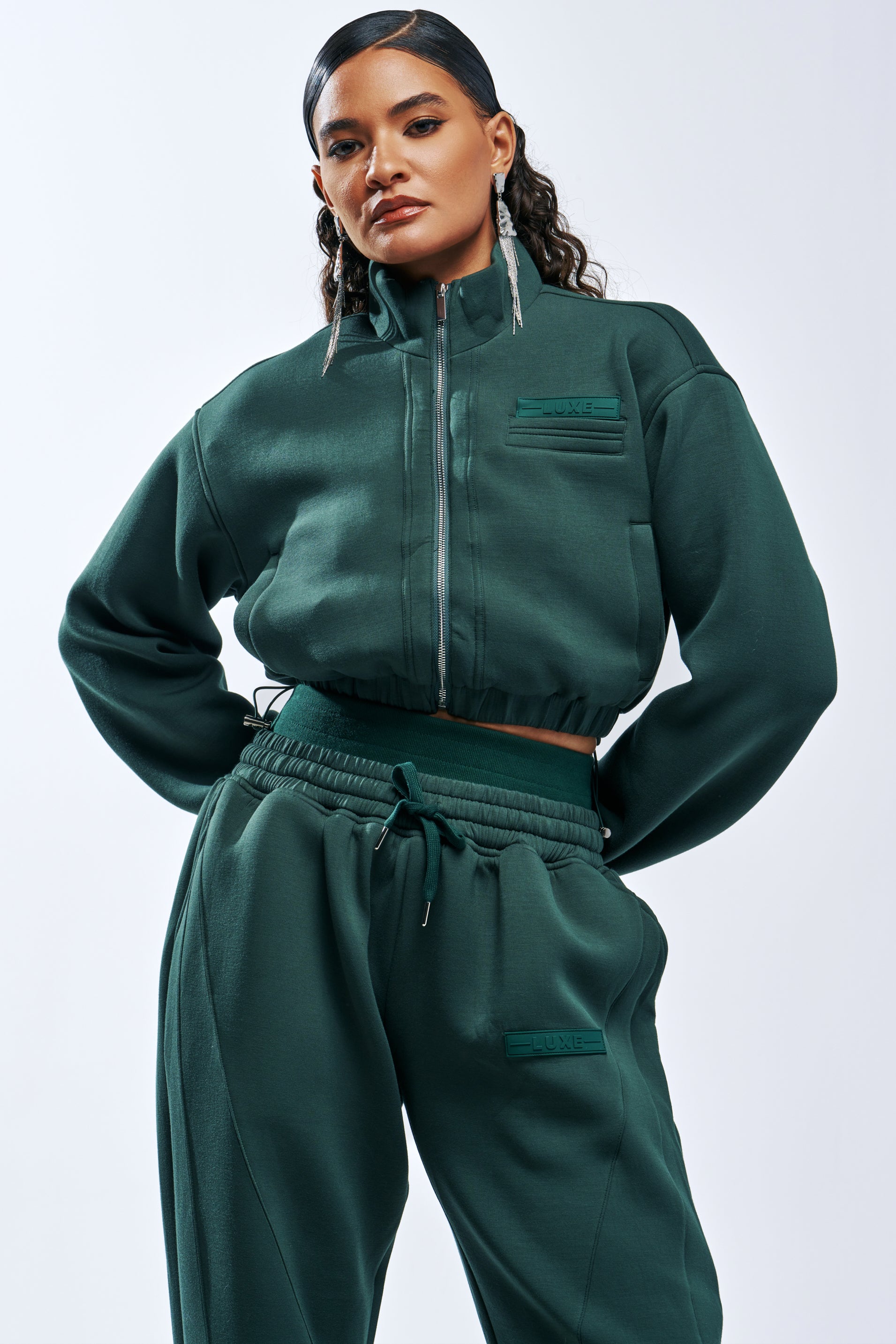A person wearing a forest green PAXTON LUXE EVERDAY ICON ZIP UP SWEATSHIRT and matching high-waisted sweatpants as an elevated loungewear set.