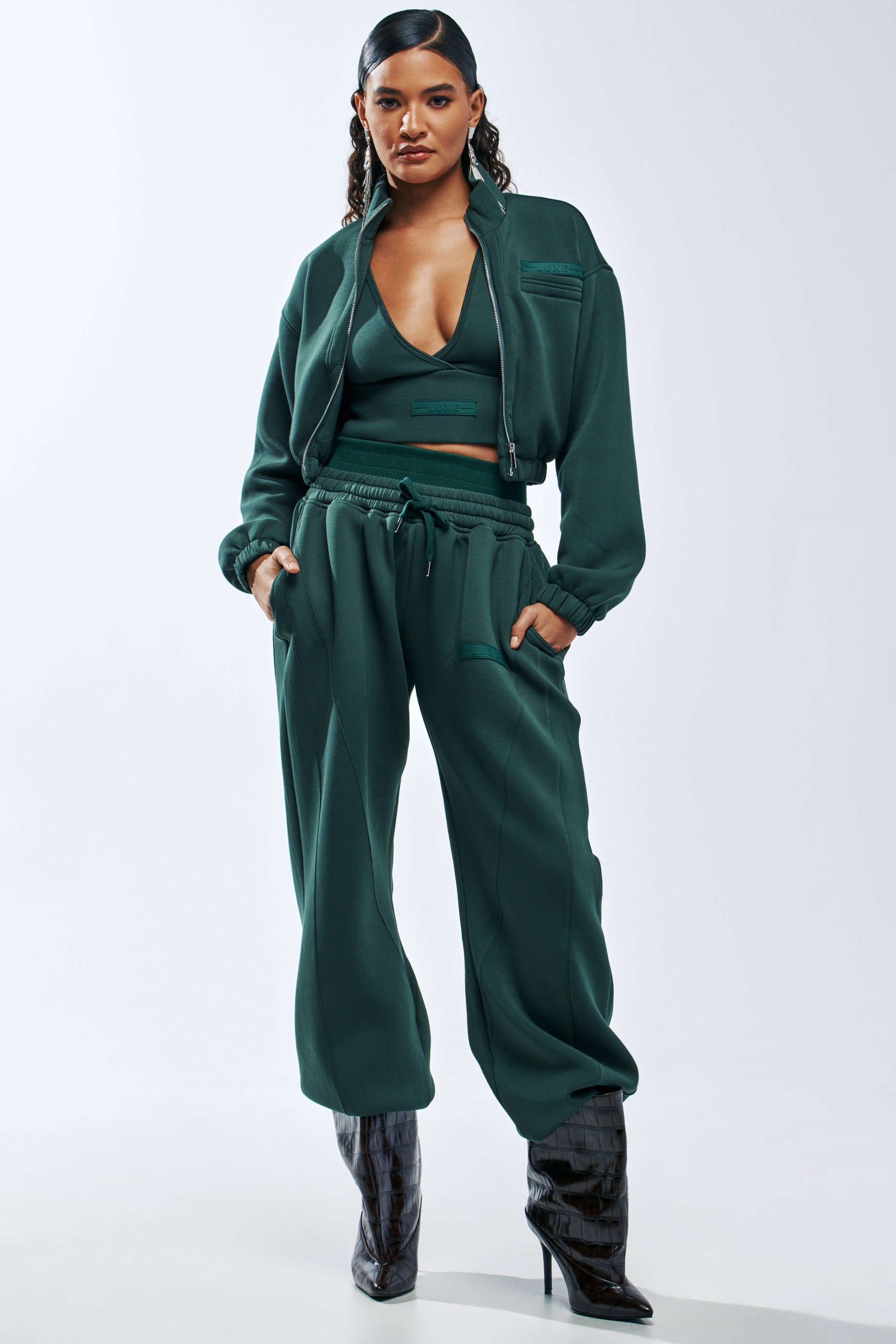 A person wears a dark green zip-up jacket, matching crop top, and PAXTON LUXE EVERYDAY ICON JOGGER wide-leg joggers with adjustable toggle hems and side pockets, paired with dark heeled boots.