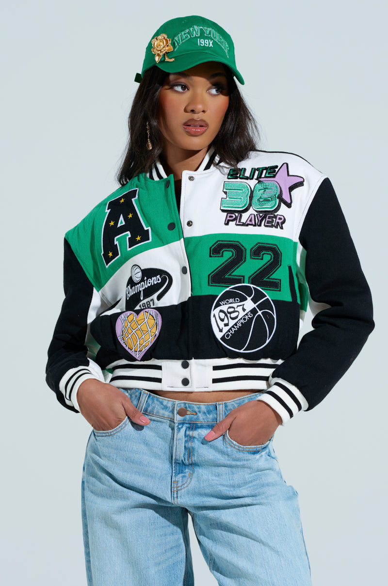 Wearing the MY RULES EMBROIDERED BOMBER featuring a large "A," basketball motifs, and numbers, paired with light blue jeans and a uniquely designed green cap against a minimalist light gray backdrop emphasizes the striking color block design.