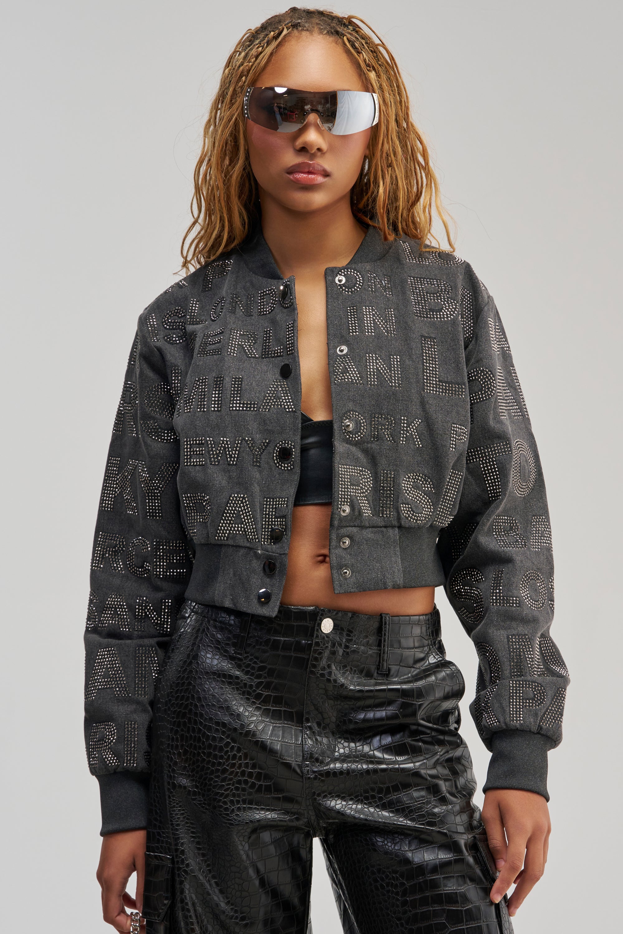 Wearing the CATCHING FLIGHTS RHINESTONE BOMBER—a black studded jacket with bold lettering—a woman with braided hair and large sunglasses pairs it with a black crop top and shiny black pants, perfectly embodying streetwear style. She stands facing forward against a plain background, her expression neutral.