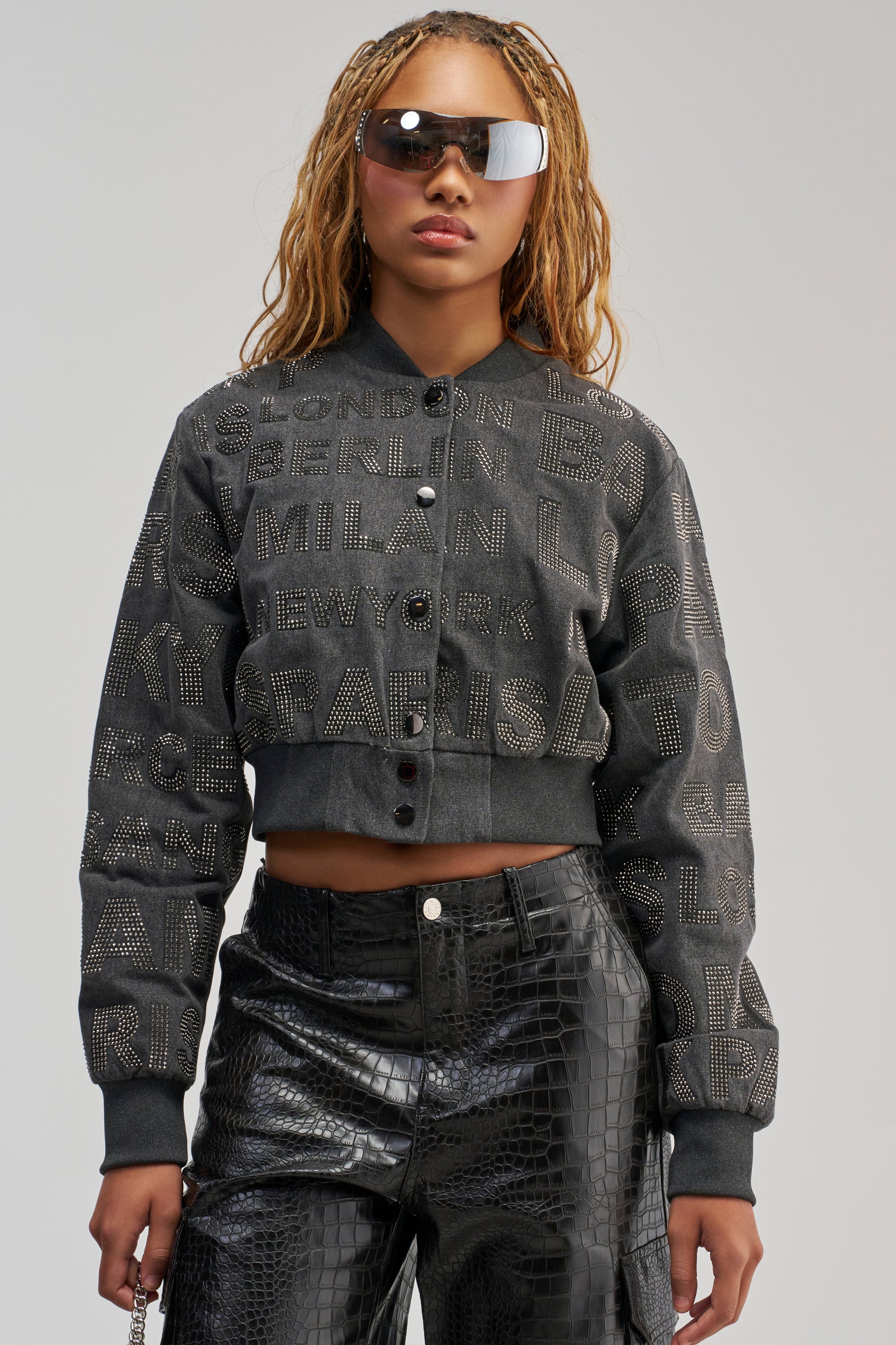 A woman wearing sunglasses and the CATCHING FLIGHTS RHINESTONE BOMBER—a textured black jacket featuring raised city names in a streetwear-inspired design—paired with shiny black pants, stands against a plain light background.
