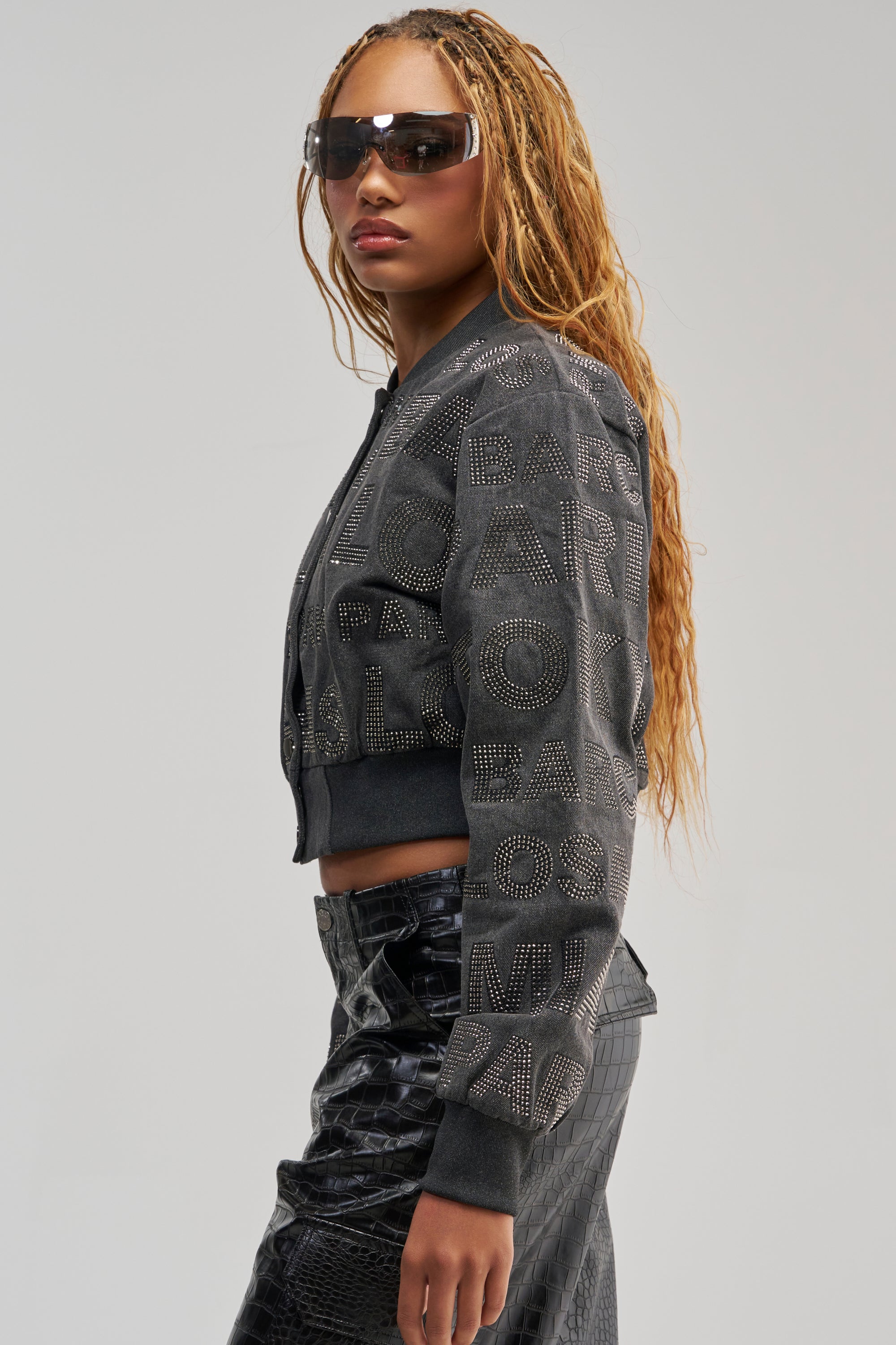 A woman with long, wavy hair wears sunglasses and the CATCHING FLIGHTS RHINESTONE BOMBER—a studded black streetwear jacket with bold lettering—paired with a matching textured skirt, standing sideways against a plain background.