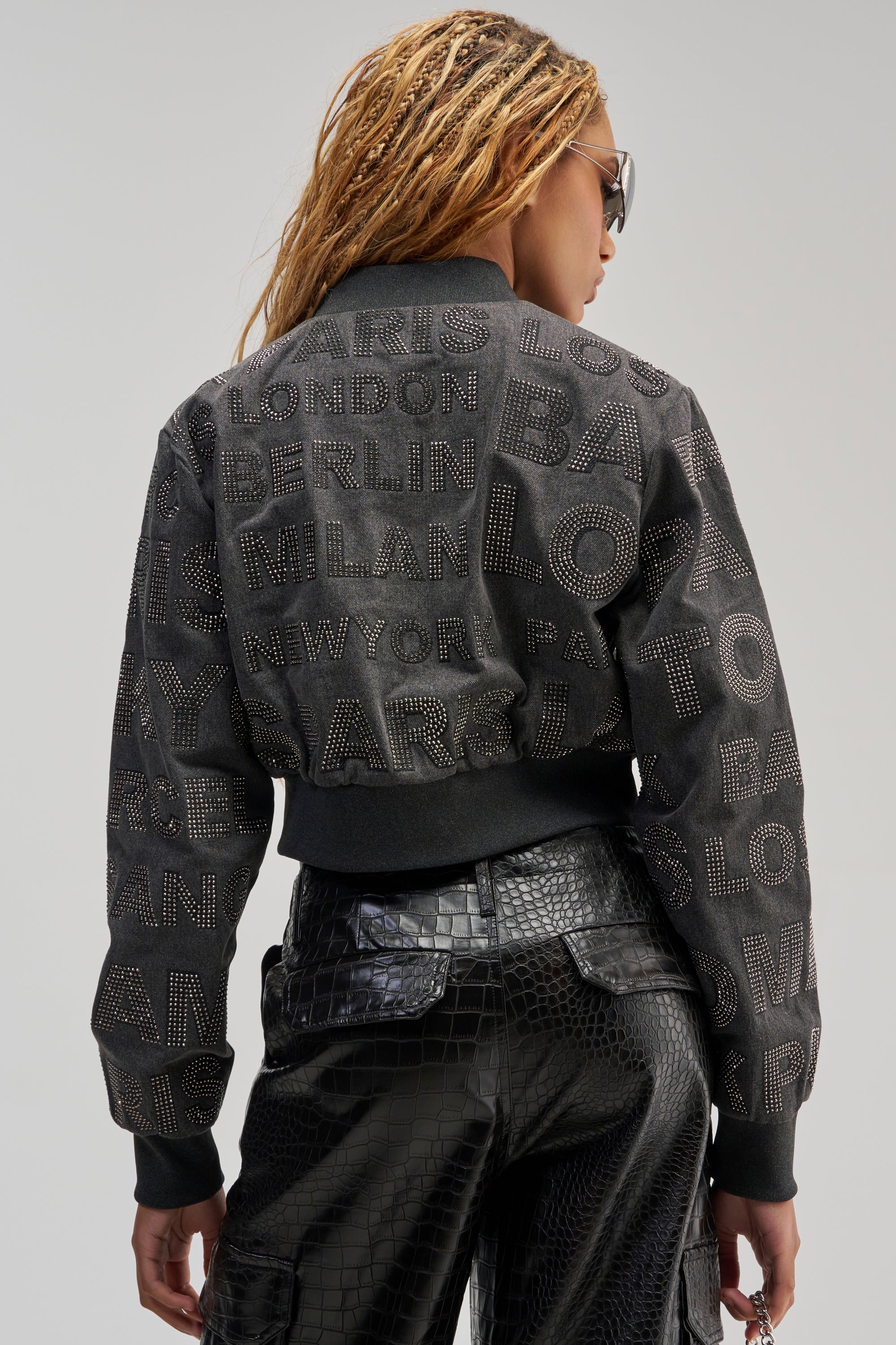 A person with long braids and sunglasses showcases the CATCHING FLIGHTS RHINESTONE BOMBER—a studded black jacket adorned with city names that channels an urban streetwear vibe—styled with shiny black crocodile-patterned pants, photographed from behind against a plain light backdrop.