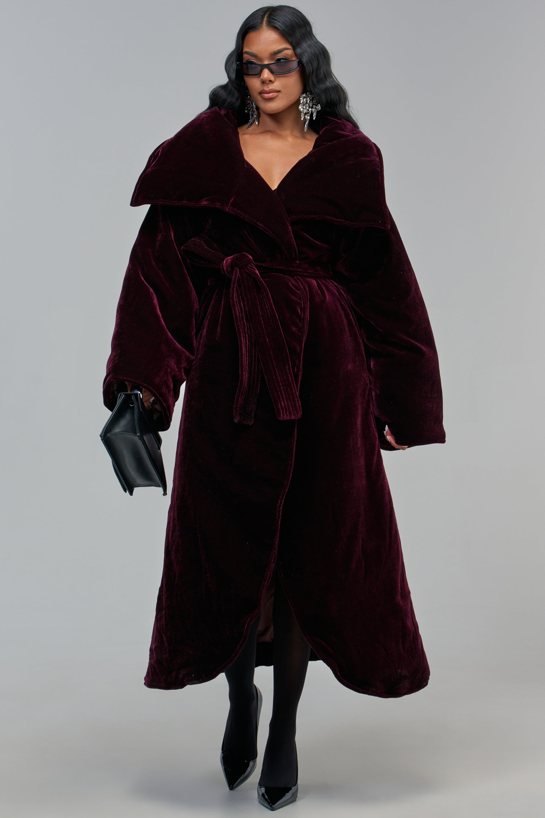 A person wears the CHAMPAGNE PLEASE VELVET TRENCH in a plum, longline, wrap-front silhouette, styled with dark sunglasses, statement earrings, and a black handbag.