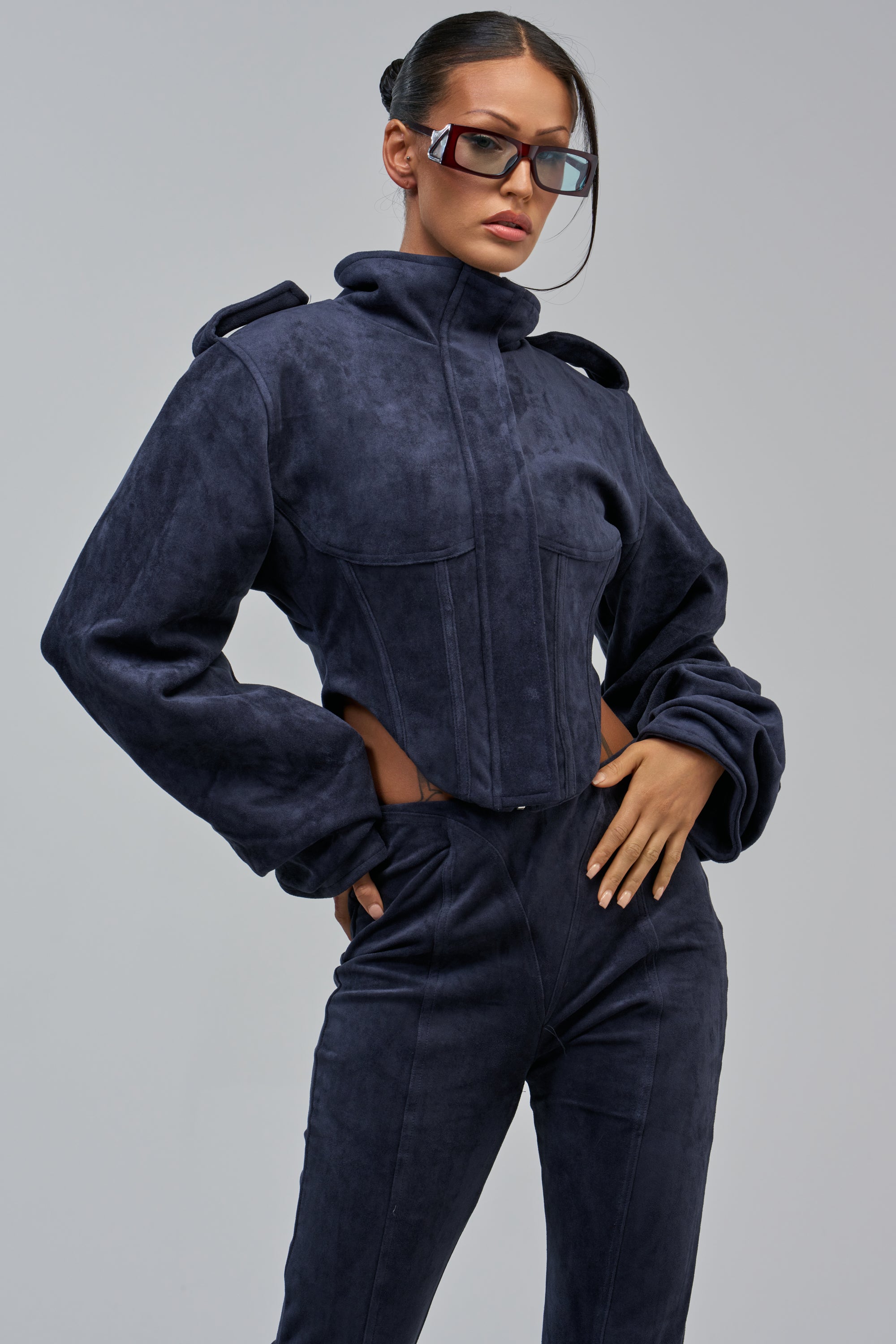 Woman wearing the KAIA FAUX SUEDE MOTO IN NAVY with a corseted waist poses confidently, hand on hip, in square sunglasses and a sleek bun against a plain gray background.