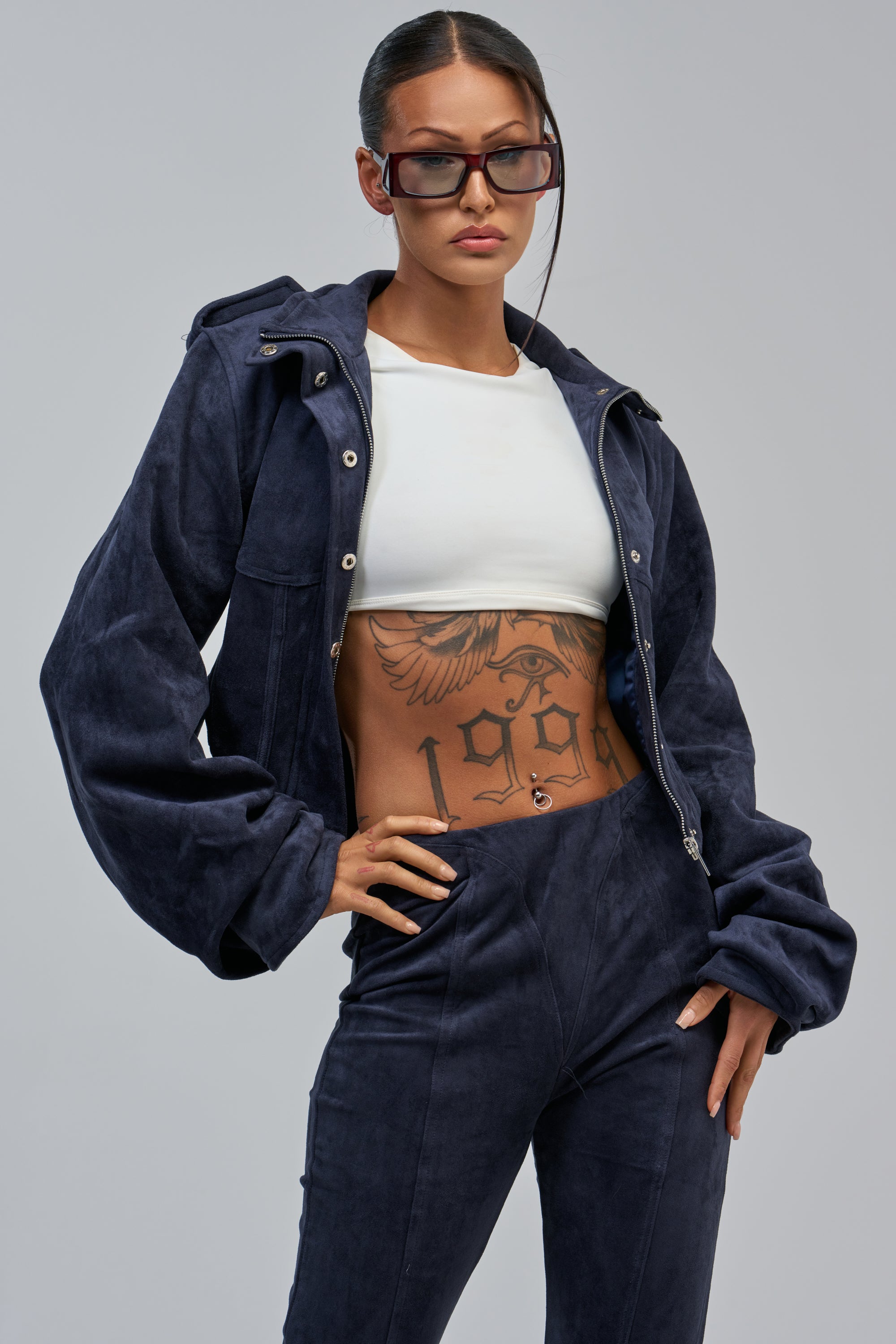 Woman wears sunglasses, a cropped white top, and the KAIA FAUX SUEDE MOTO IN NAVY with pants. Her corseted waist and “1998” eagle tattoo stand out as she poses confidently against a plain light background.