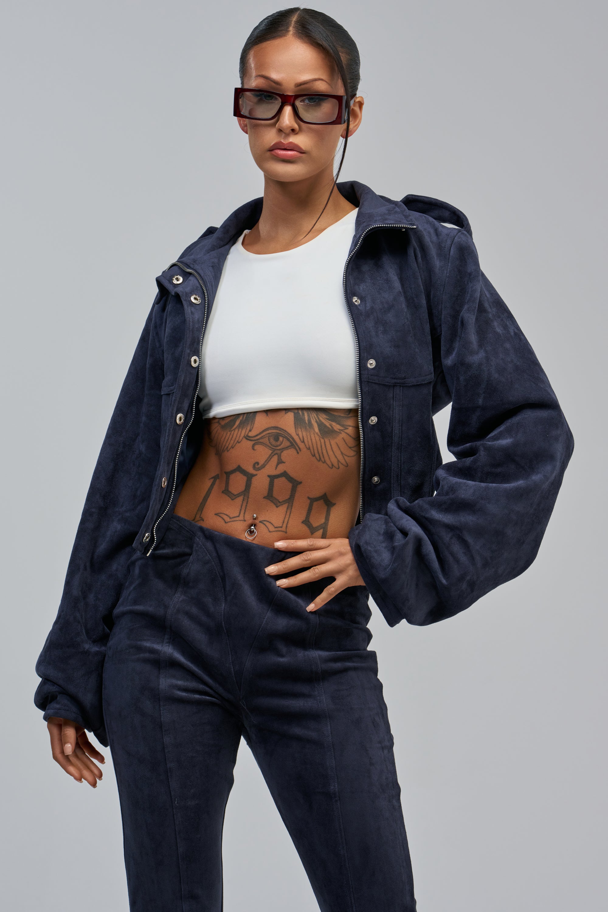 Wearing the KAIA FAUX SUEDE MOTO IN NAVY and matching pants with a white crop top and large sunglasses, this look features bold tattoos—perfect for fall layering.
