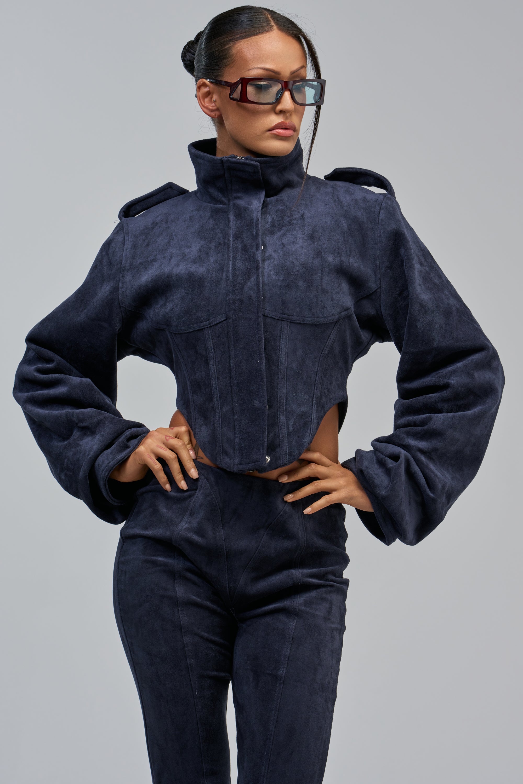 A woman poses in the KAIA FAUX SUEDE MOTO IN NAVY with matching pants and large rectangular sunglasses, her hair tied back, creating an effortlessly layered fall look against a light gray background.