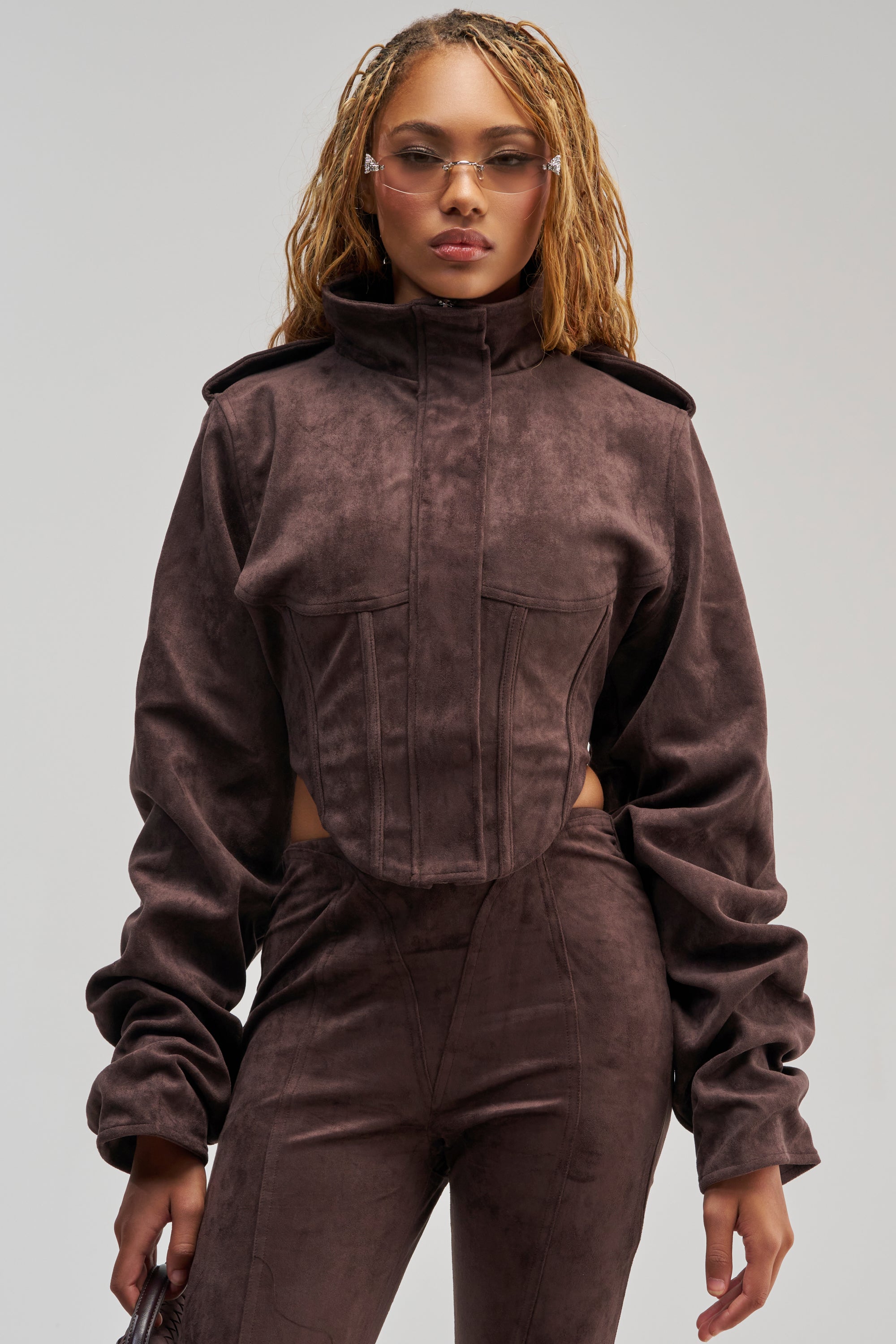 A person stands facing forward against a plain background, wearing the KAIA FAUX SUEDE MOTO jacket with structured shoulders and matching pants, accessorized with a small bag, clear glasses, and long styled hair.