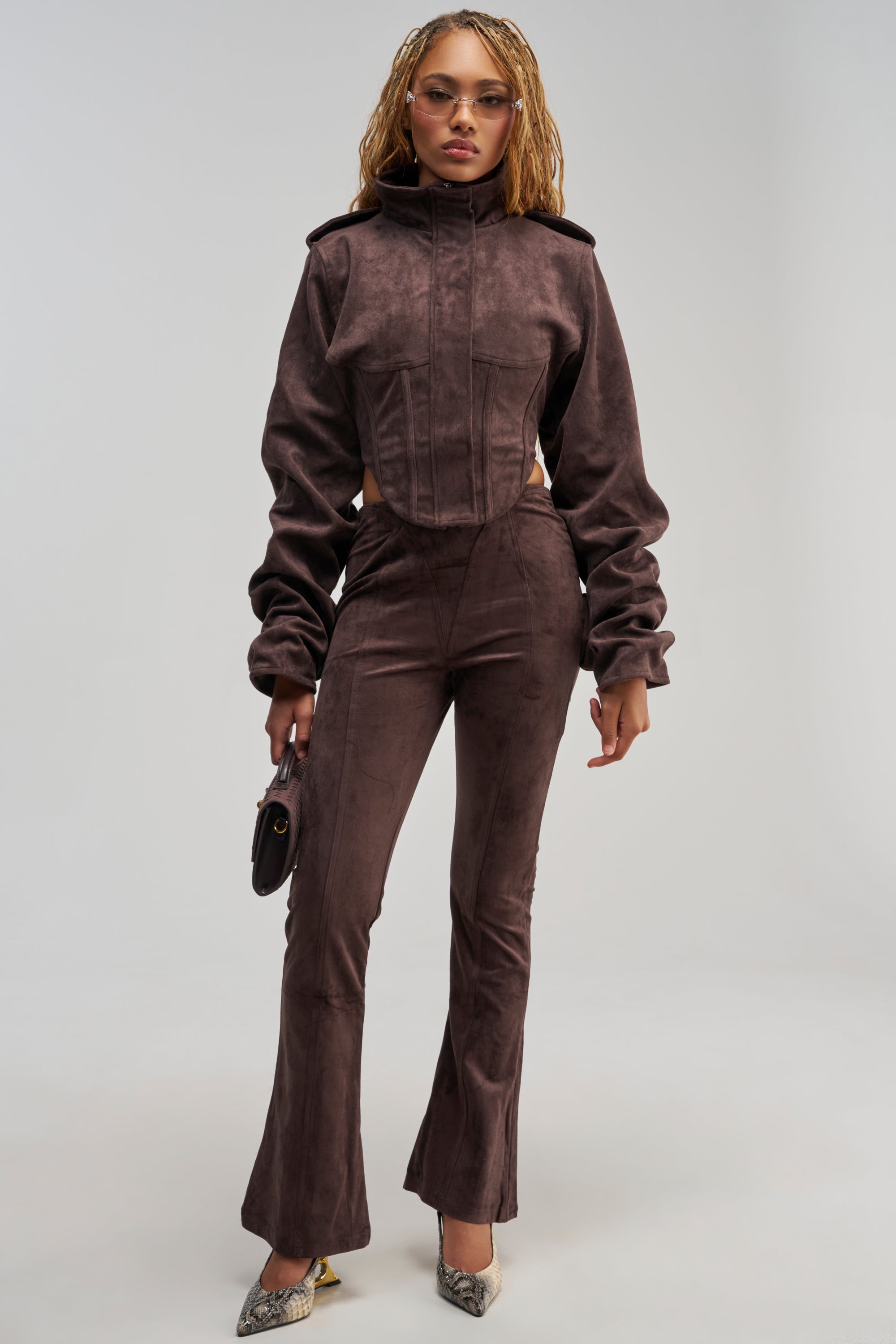 A woman stands against a plain light background wearing the KAIA FAUX SUEDE FLARE LEGGING in brown, paired with a fitted dark brown suede jacket, accessorized with round glasses, a small purse, and snakeskin boots.