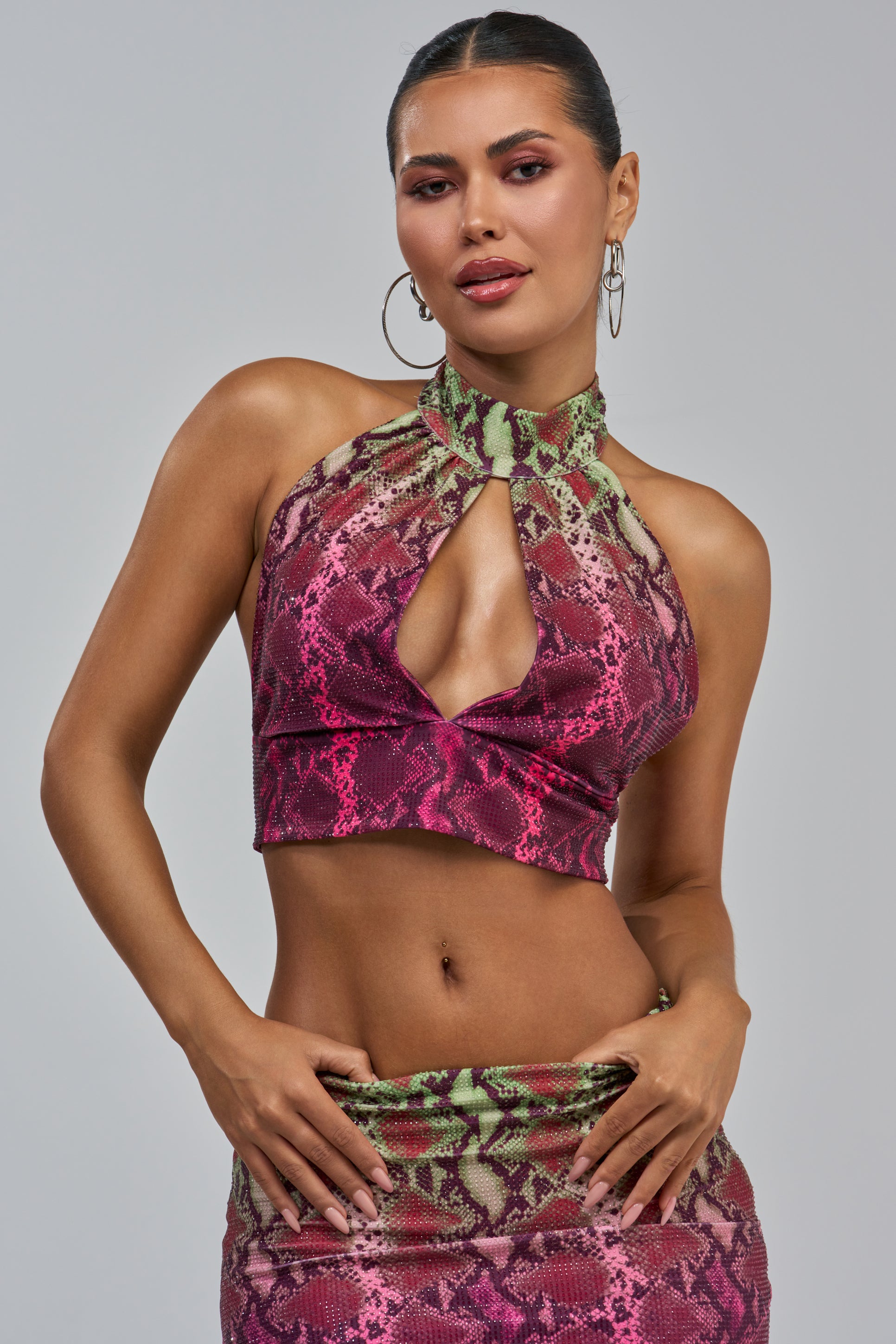 A woman poses with her hands on her hips against a plain light gray background, wearing the YOU LITTLE SEXY SNAKE BLOUSE—a pink and green rhinestone snakeskin halter crop top styled with a matching skirt.