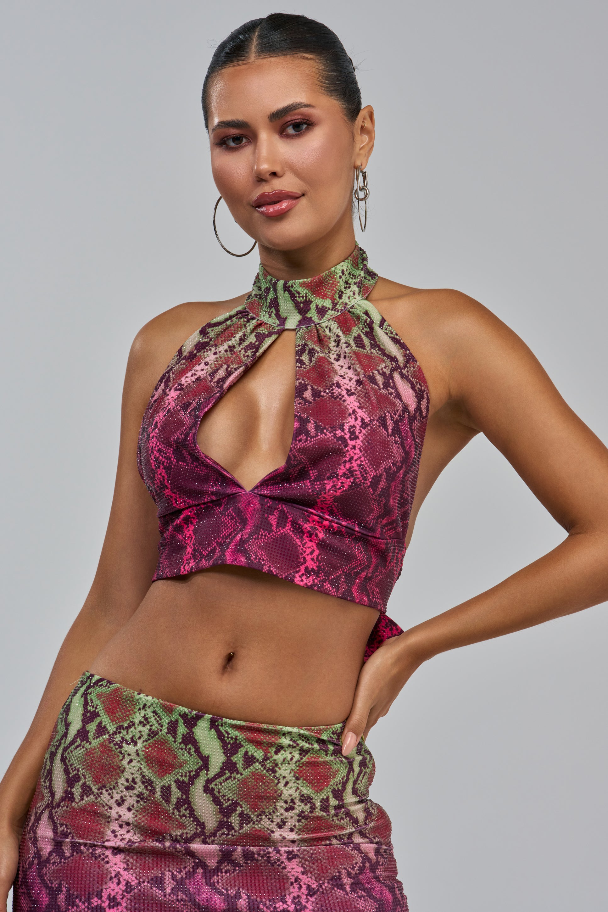 A woman poses confidently, wearing the YOU LITTLE SEXY SNAKE BLOUSE—a sleeveless, high-neck party top featuring a vibrant snake print in shades of pink, green, and purple. She pairs it with a matching skirt, accessorizes with hoop earrings, and has her hair pulled back.