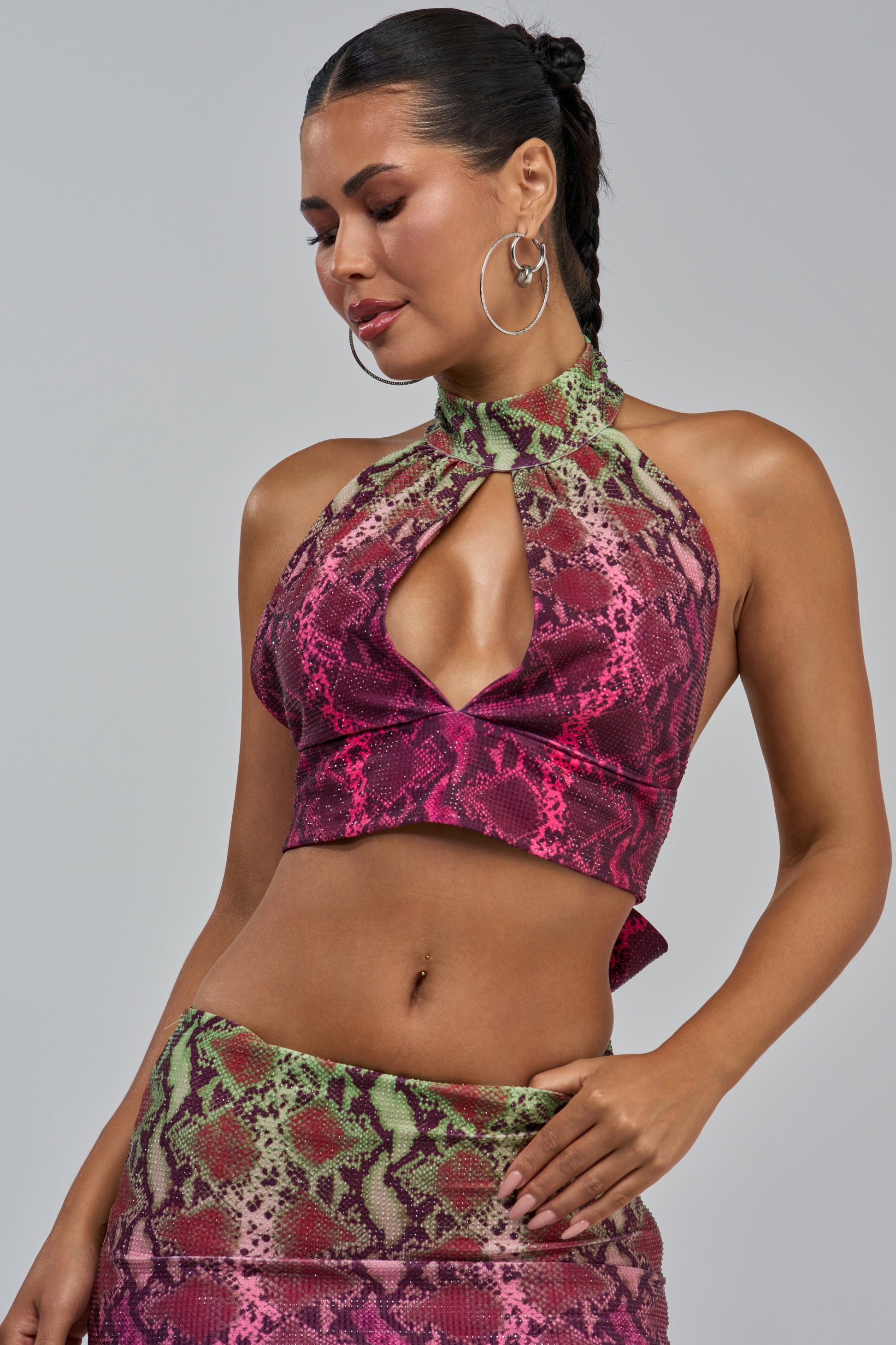 A woman poses against a plain gray background wearing the YOU LITTLE SEXY SNAKE BLOUSE, a colorful rhinestone snakeskin top styled as a halter and skirt set with a keyhole cutout. She accessorizes with large hoop earrings and wears her hair in braids.