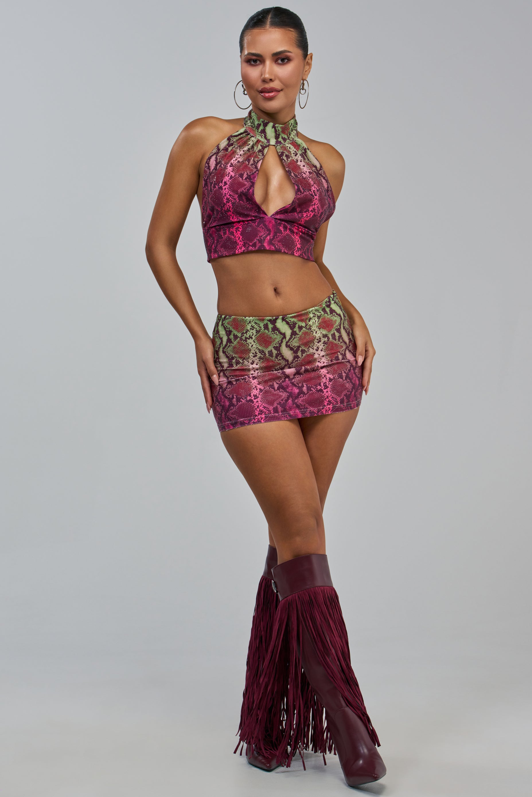 Wearing the YOU LITTLE SEXY SNAKE MINI SKIRT in a pink and green snakeskin pattern, paired with a matching halter crop top, burgundy fringe boots, hoop earrings, and her hair pulled back.