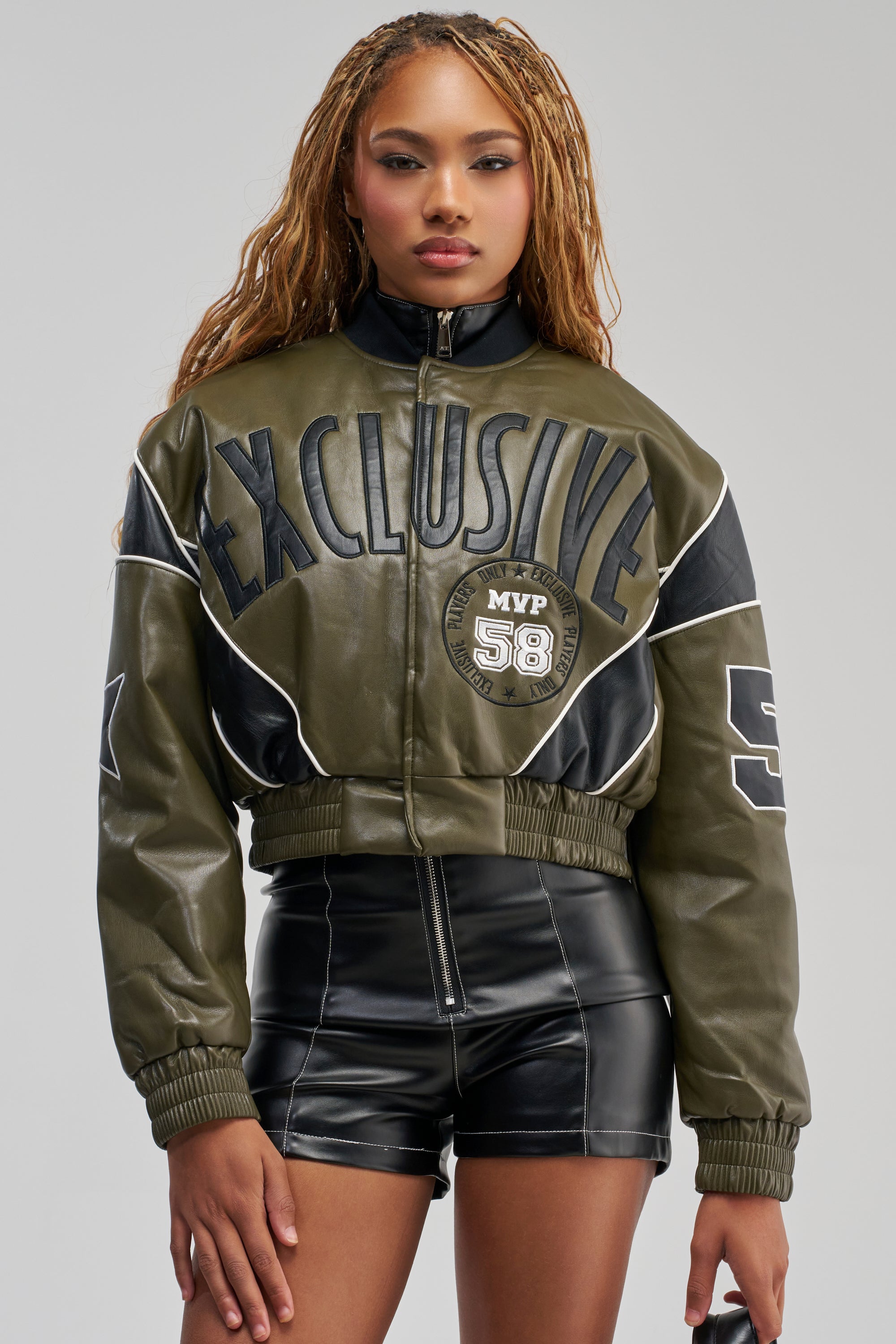 Wearing the BETTER BELIEVE VARSITY BOMBER—an oversized, cropped olive green faux leather jacket with “EXCLUSIVE” and “MVP 58” detailing—and paired with high-waisted black leather shorts, a person stands facing forward against a plain background, showcasing a modern streetwear look.