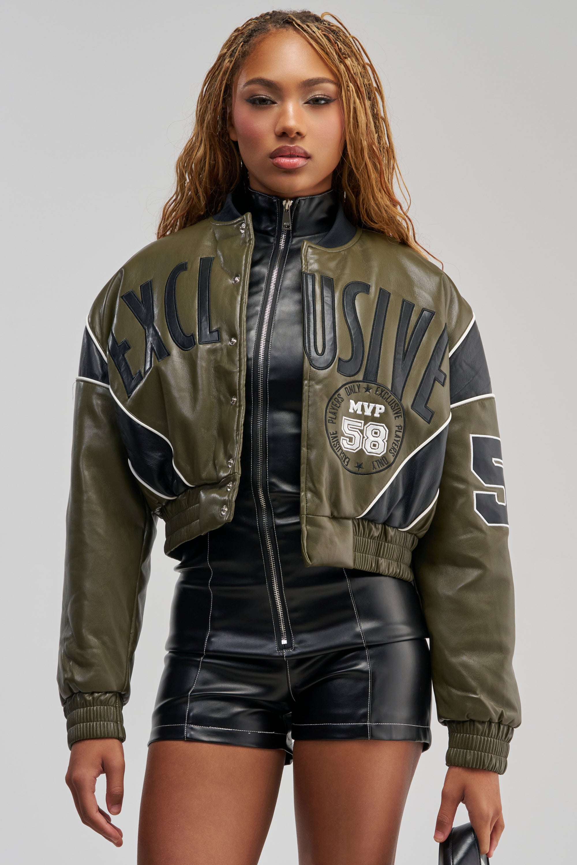 A woman with long braided hair models the BETTER BELIEVE VARSITY BOMBER in green and black over a sleek black faux leather jumpsuit, highlighting bold streetwear layering. Standing against a neutral background, she faces forward while holding an object in her right hand.