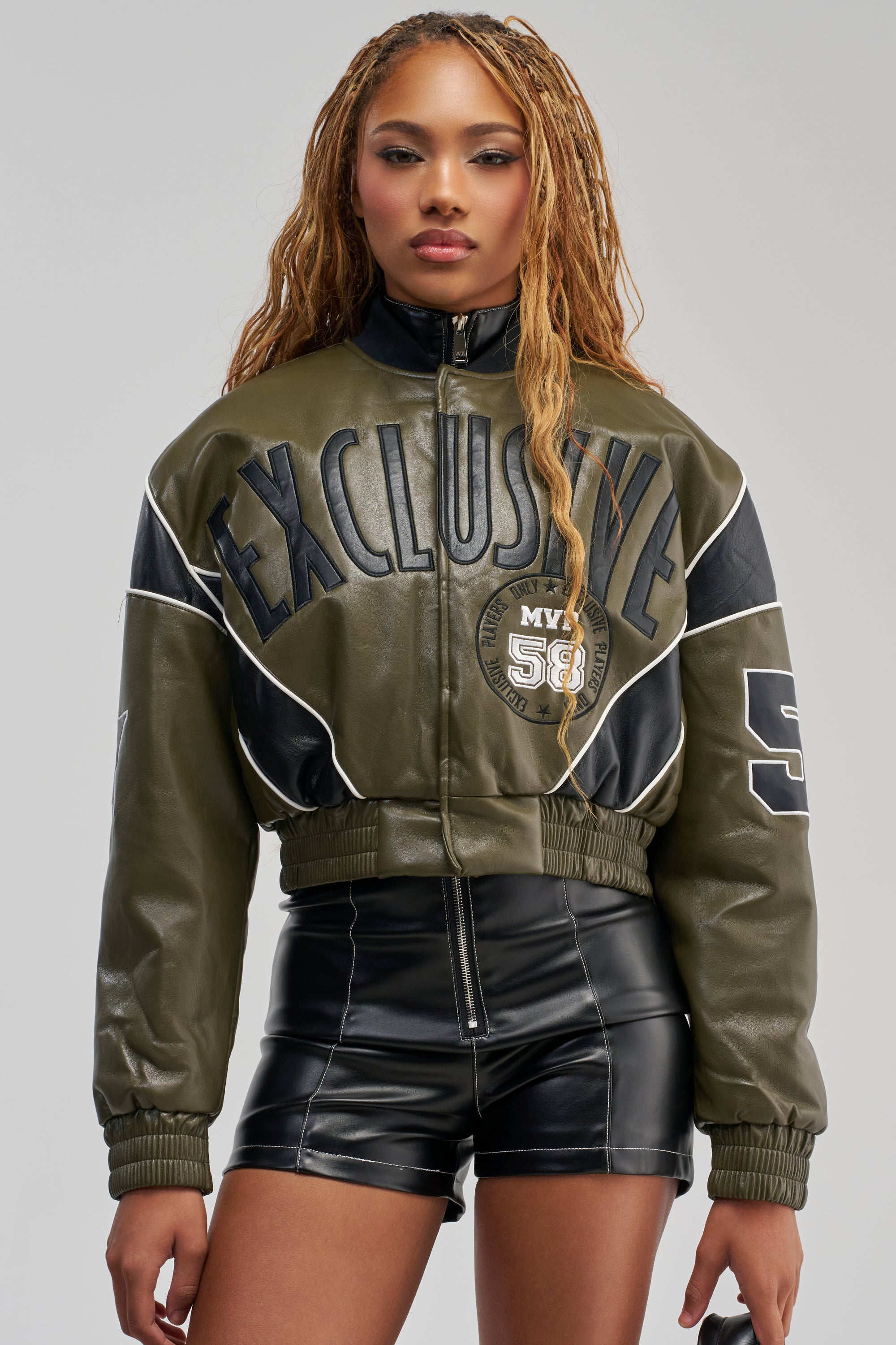 A woman with long braids wears the BETTER BELIEVE VARSITY BOMBER—an olive green faux leather streetwear jacket with an oversized, cropped fit and “EXCLUSIVE” lettering—paired with black high-waisted leather shorts. She stands facing forward against a plain light background.