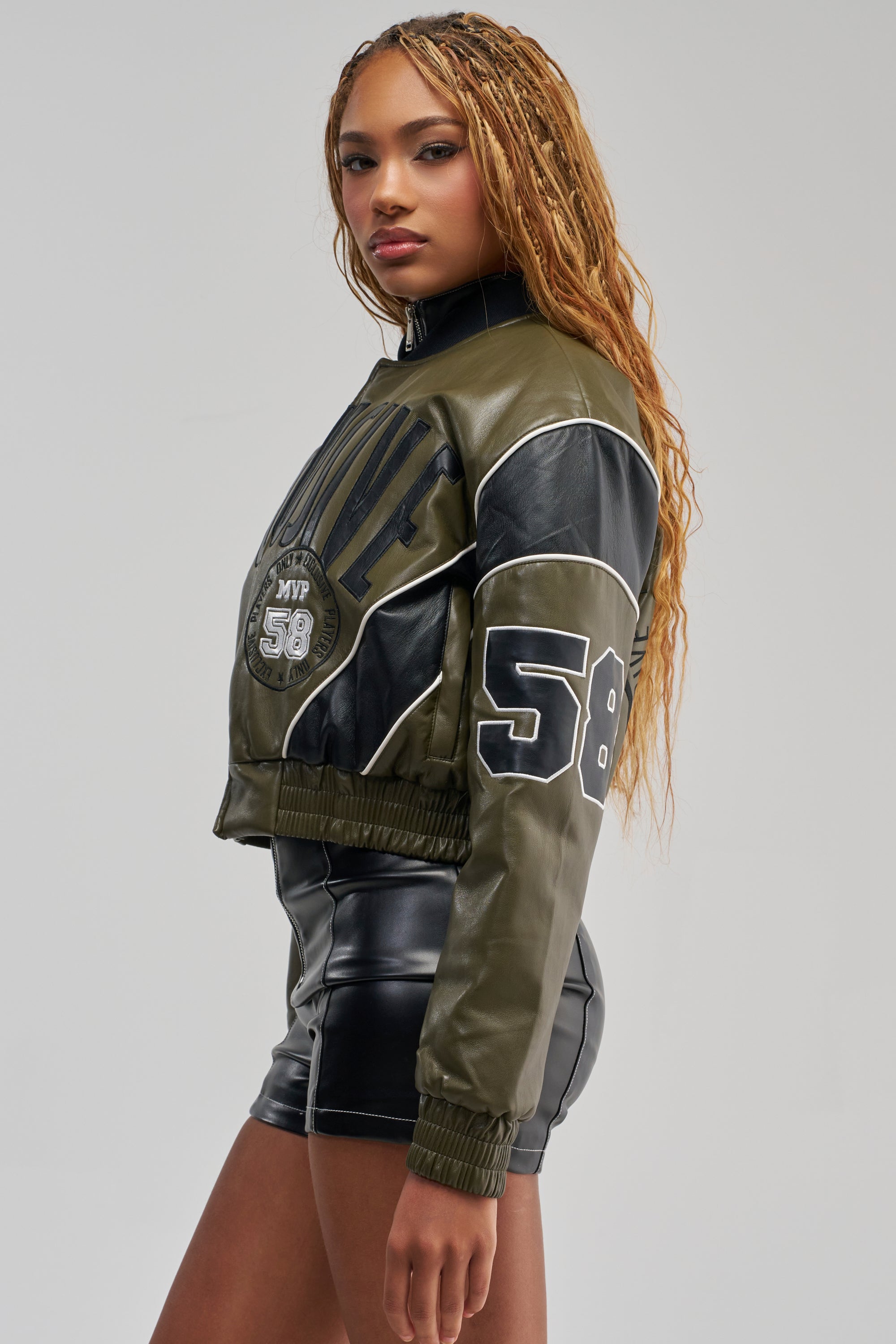 A woman with long braided hair poses against a plain gray background, wearing the BETTER BELIEVE VARSITY BOMBER in olive green faux leather—an oversized, cropped varsity jacket with “58” on the sleeve—paired with black leather shorts for a bold streetwear look.