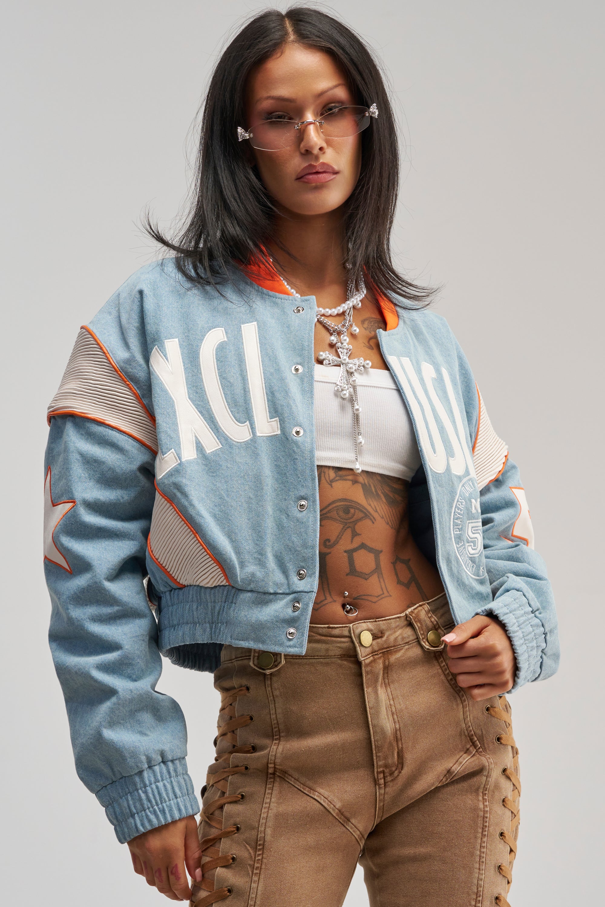 Dressed in the INVITE ONLY DENIM VARSITY BOMBER, a woman with straight dark hair pairs her look with tan lace-up pants, a white crop top, layered necklaces, and pink sunglasses as she stands confidently against a plain background.