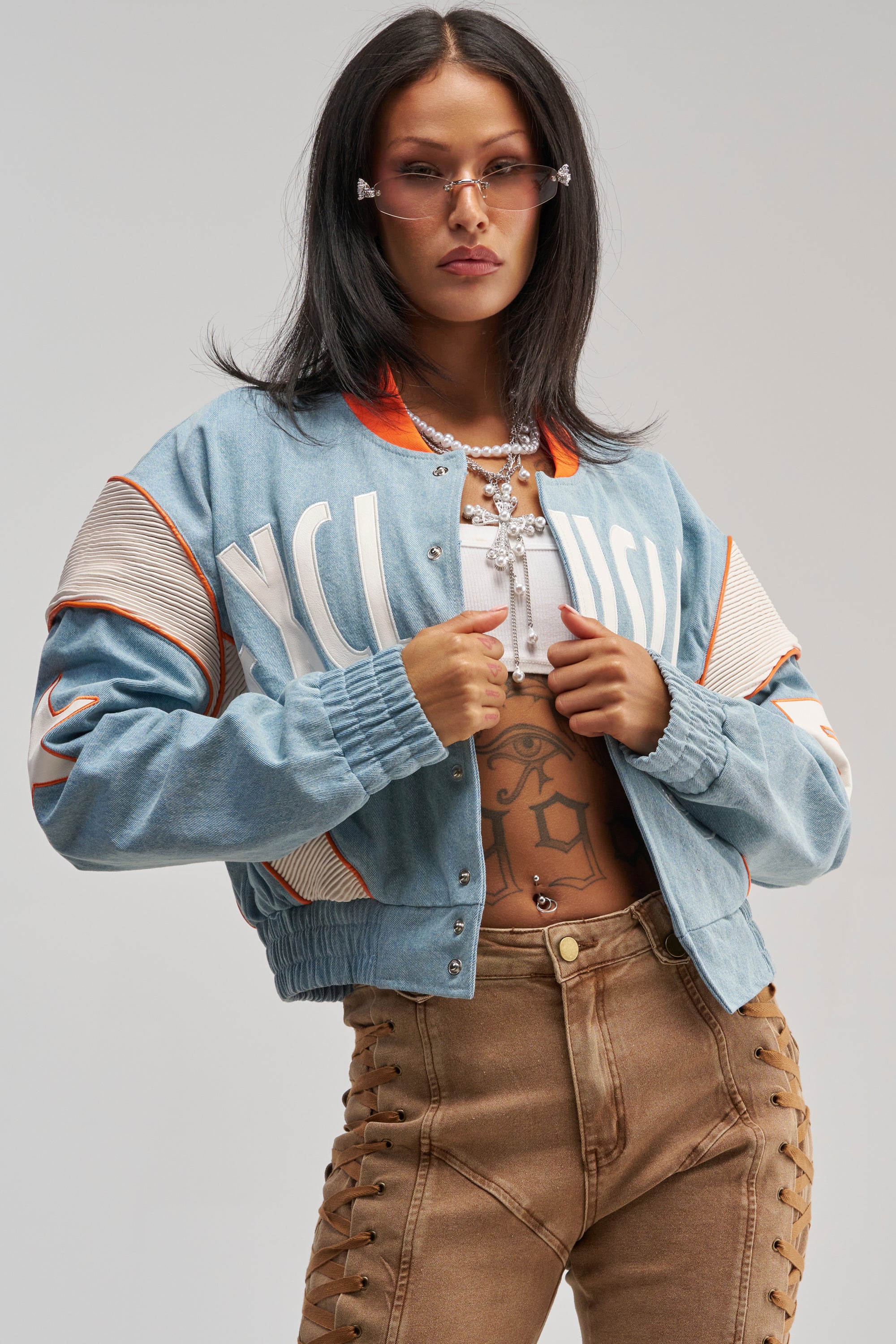 A woman wearing the INVITE ONLY DENIM VARSITY BOMBER, tan lace-up pants, sunglasses, and layered necklaces poses confidently with her hands on her jacket.