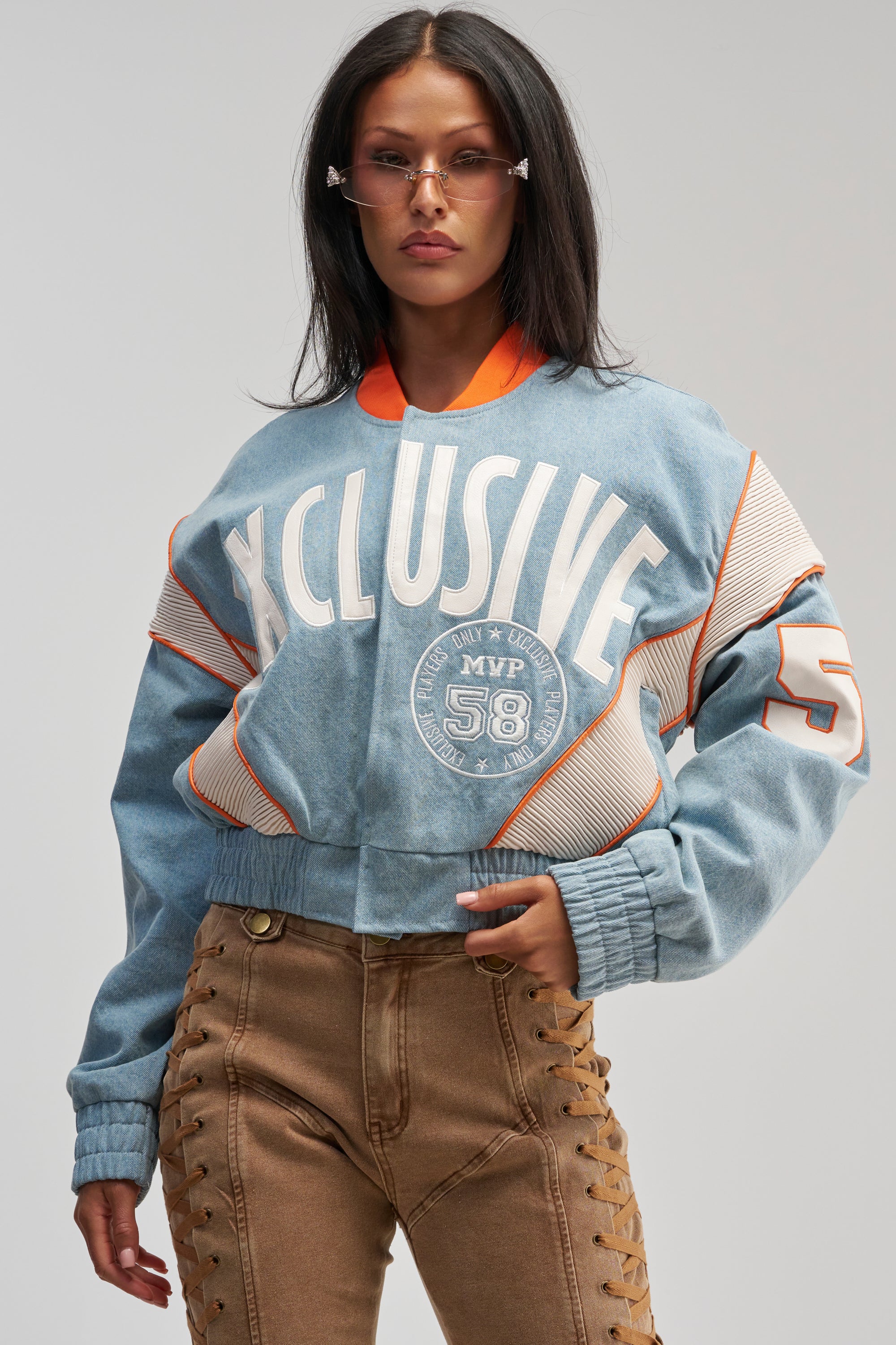 A woman poses confidently while looking at the camera, wearing the INVITE ONLY DENIM VARSITY BOMBER— a blue jacket with orange accents— paired with brown lace-up pants. She has long dark hair and is accessorized with clear-framed sunglasses.