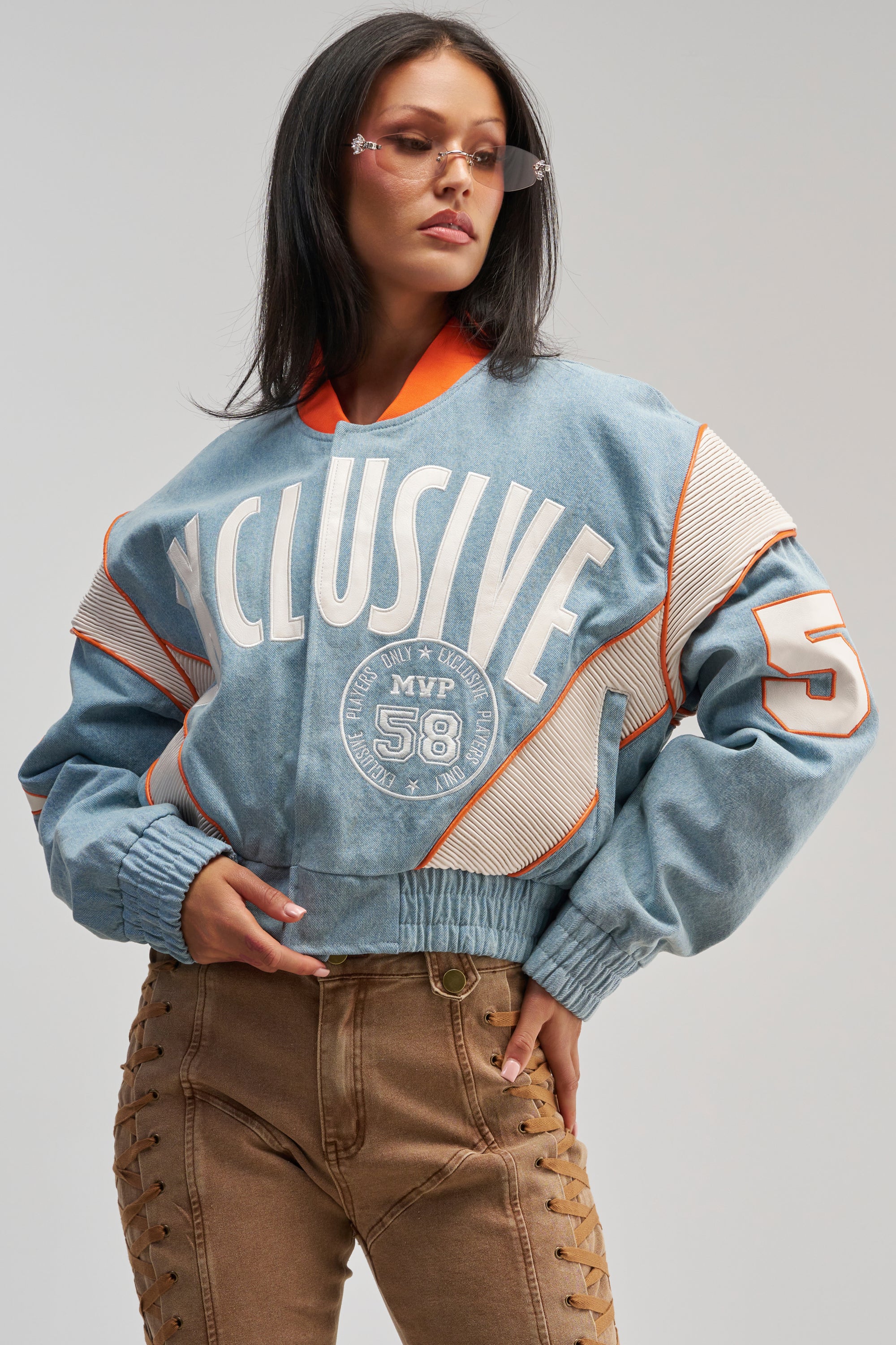 A woman stands confidently against a plain background, wearing tinted glasses and high-waisted brown lace-up pants, her blue oversized “XCLUSIVE” sweatshirt with orange accents effortlessly layered beneath the INVITE ONLY DENIM VARSITY BOMBER for an added touch of streetwear flair.