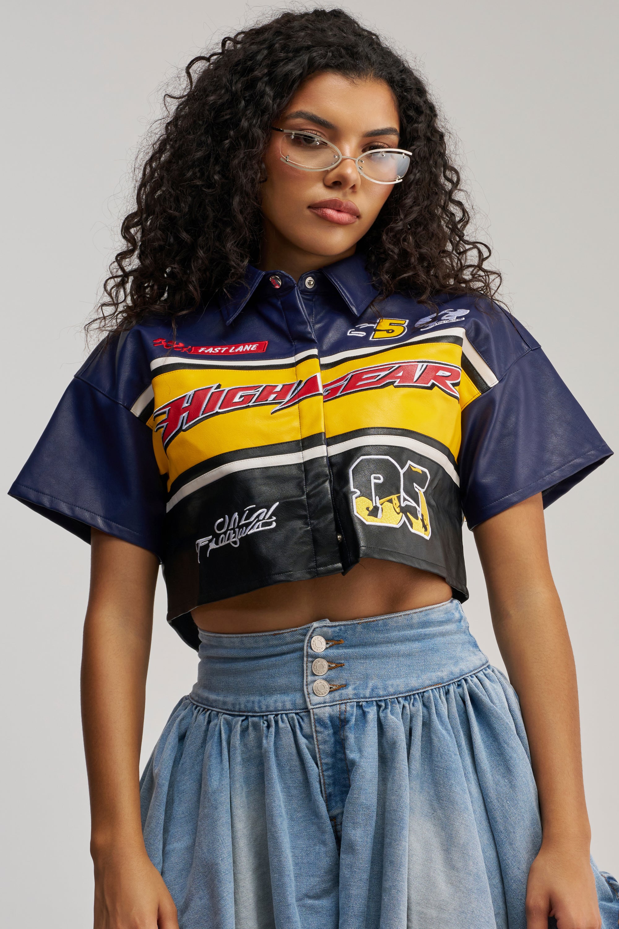 A woman with curly hair and glasses is wearing the HIGH GEAR FAUX LEATHER SHORT SLEEVE BUTTON DOWN—a cropped, color-block racing-style shirt featuring yellow, red, and blue patterns—paired with a high-waisted button-front denim skirt, standing against a plain background.