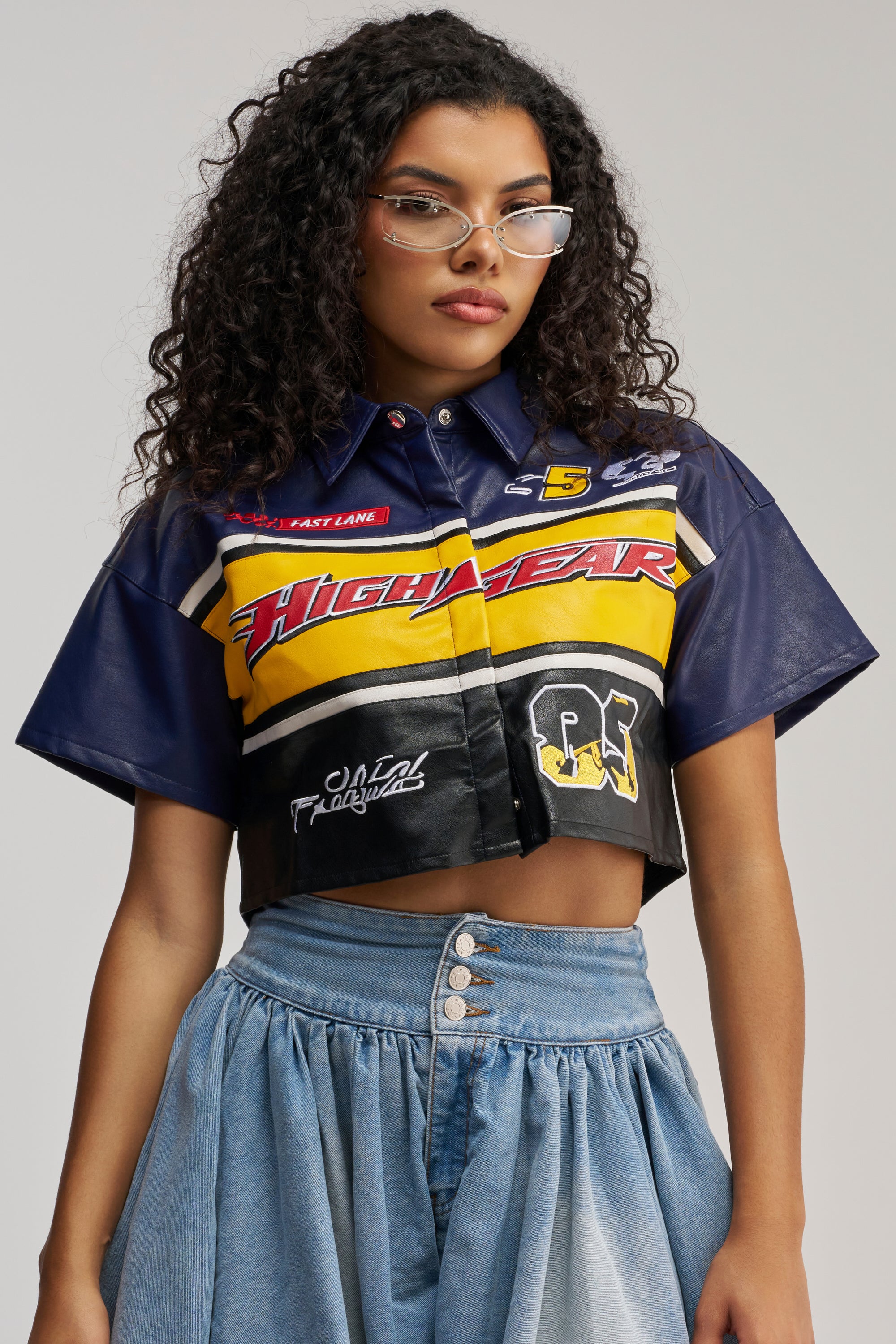 Wearing the HIGH GEAR FAUX LEATHER SHORT SLEEVE BUTTON DOWN, a woman with curly hair and cat-eye glasses poses against a plain light background, embodying effortless streetwear style. She pairs the bold button-down with high-waisted blue denim jeans for a striking look.