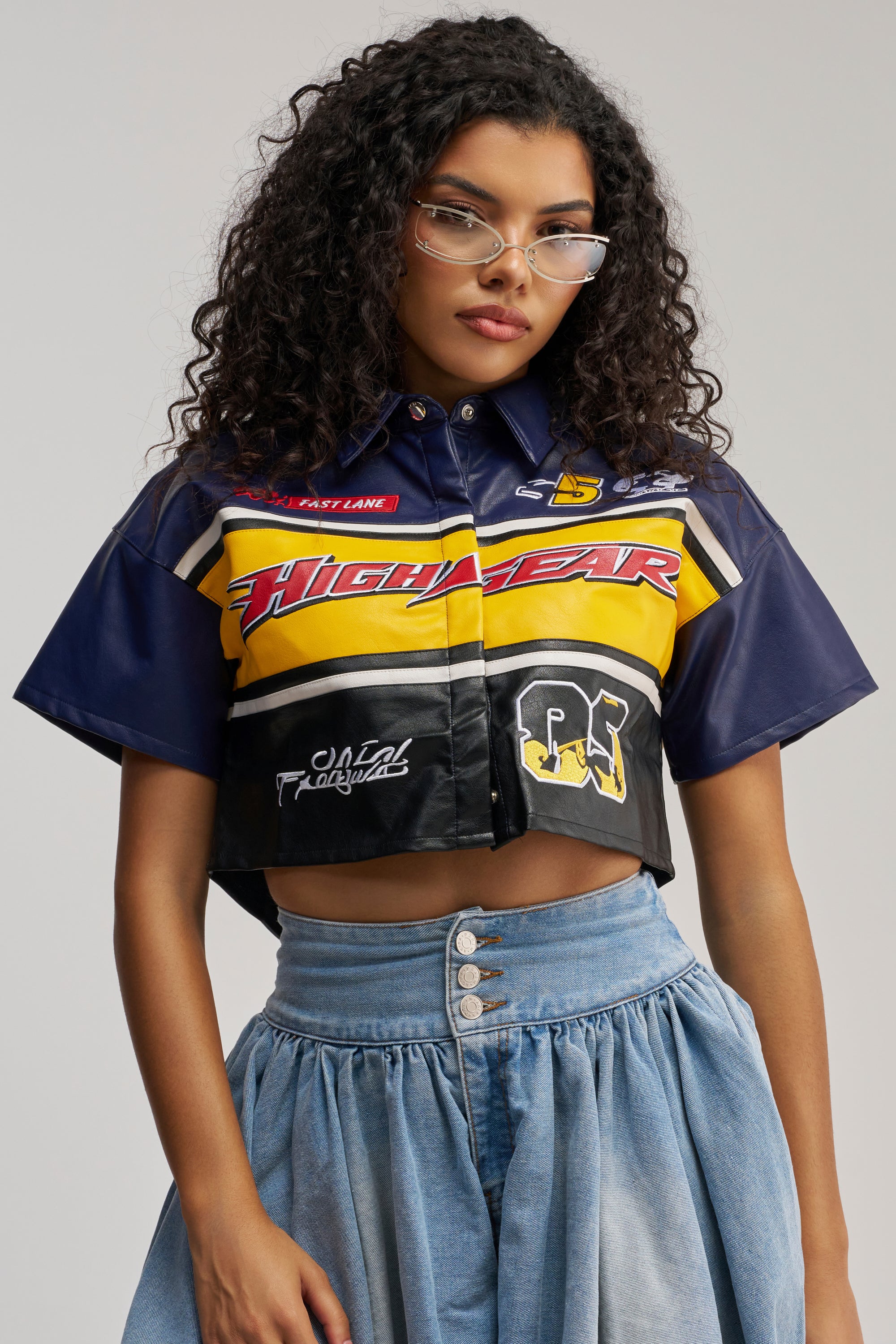 A person with curly hair and glasses wears the HIGH GEAR FAUX LEATHER SHORT SLEEVE BUTTON DOWN styled as a cropped, racing-inspired shirt with color block details, paired with a high-waisted denim skirt. The background is plain and light-colored.