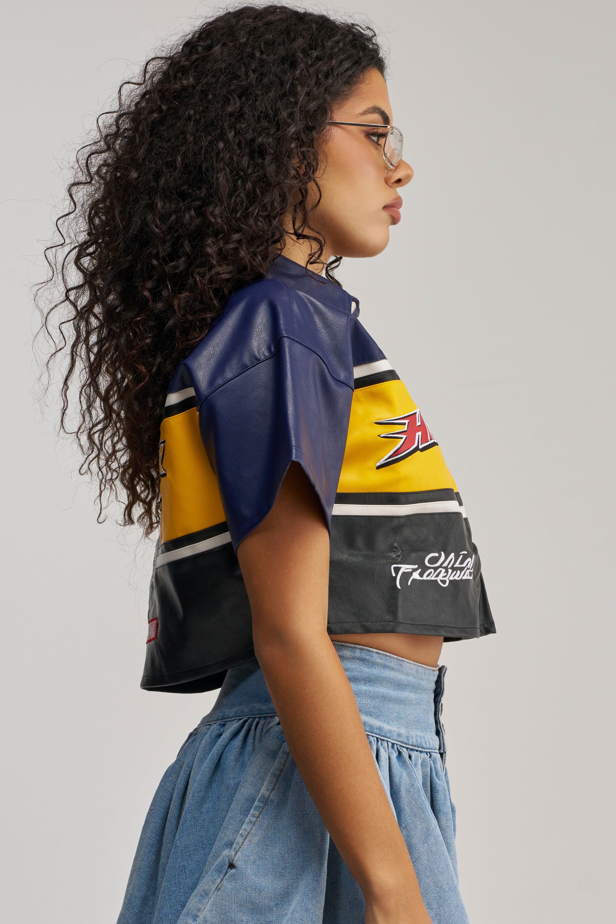 A person with curly hair and glasses wears the HIGH GEAR FAUX LEATHER SHORT SLEEVE BUTTON DOWN, styled in a streetwear-inspired look with a high-waisted denim skirt, shown in side profile against a plain background.