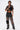 A person wearing sunglasses, a black crop top, and the STEPHANIA TRANSPARENT TROUSER IN BLACK with chunky black boots poses confidently against a plain white background.