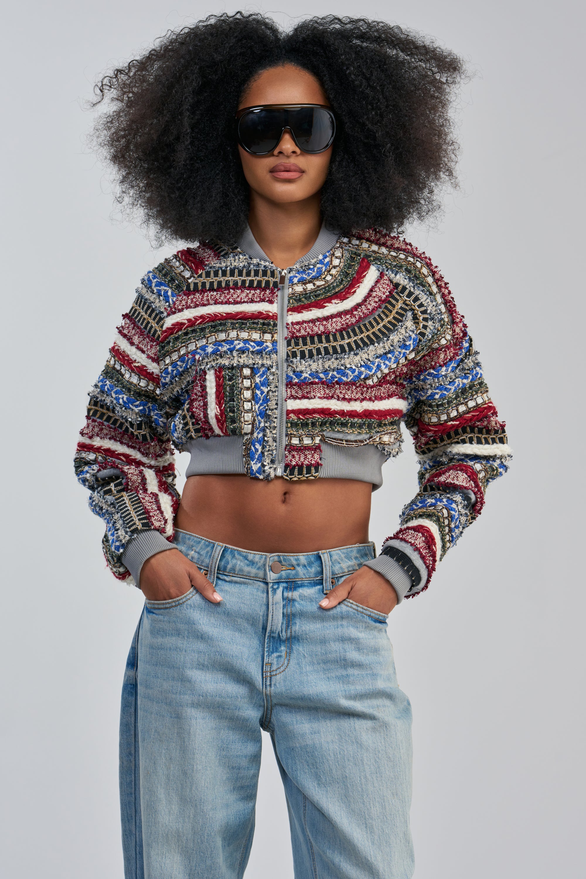 A person with voluminous curly hair wears large black sunglasses, the FAYE TEXTURED BOMBER—a colorful textured streetwear jacket—and light blue jeans, posing with hands in pockets against a plain background.