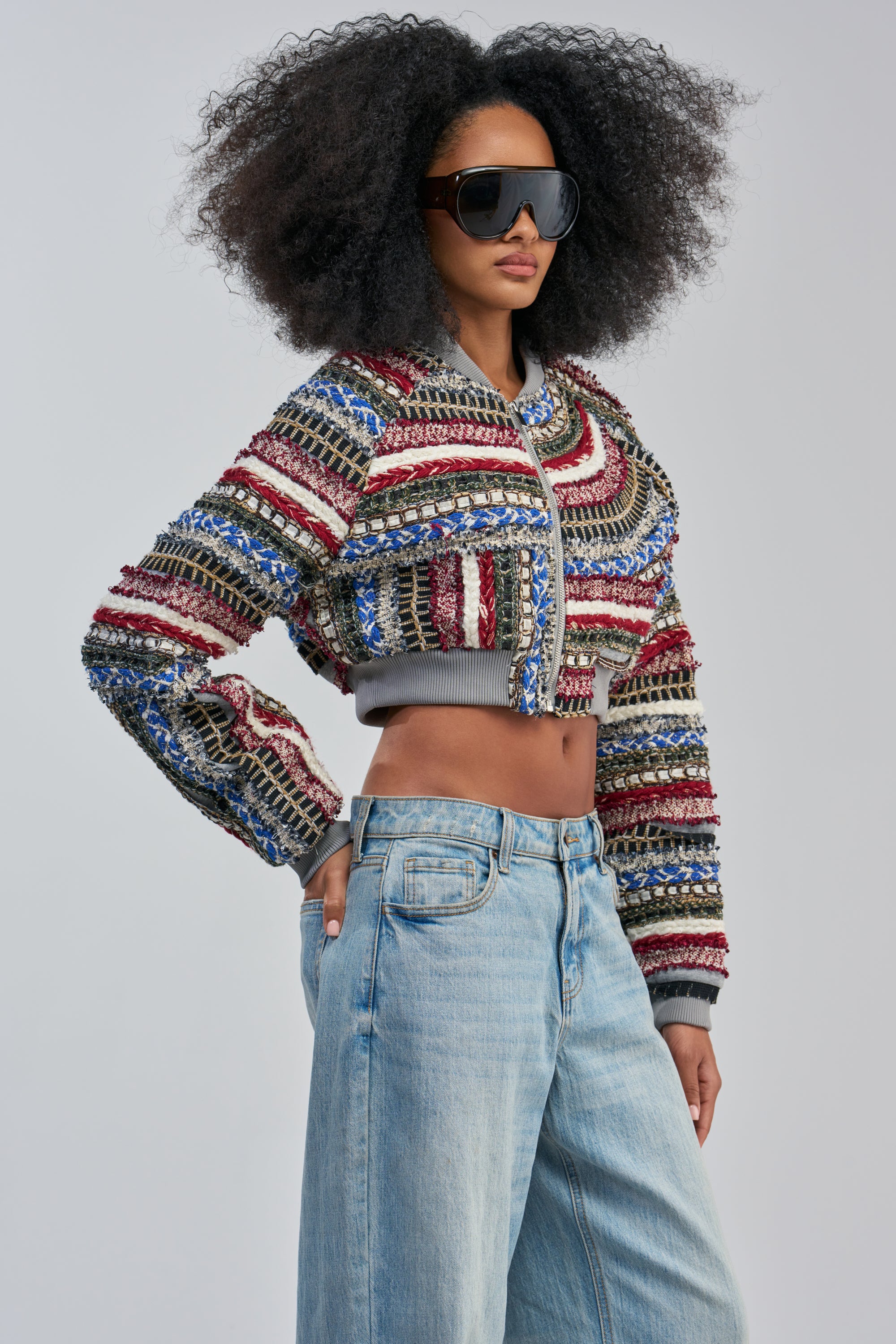 Wearing the FAYE TEXTURED BOMBER, a woman with voluminous curly hair and large sunglasses pairs her look with high-waisted light blue jeans, posing confidently against a plain background.